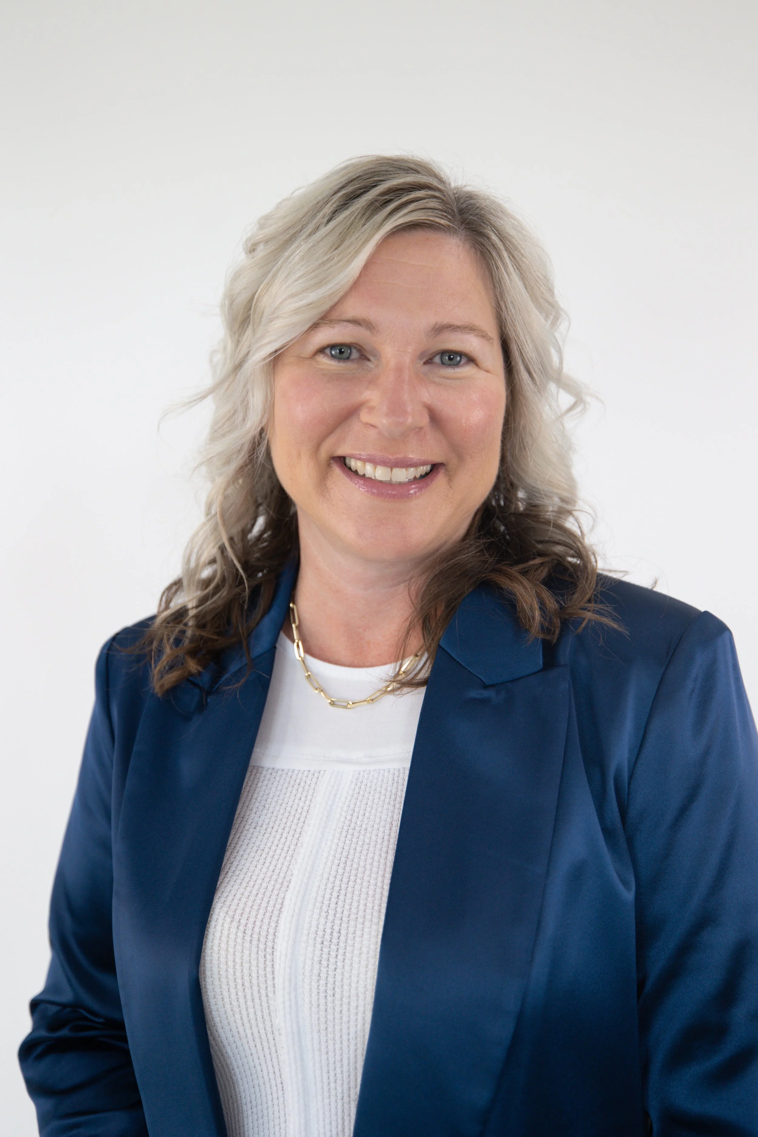 A smiling woman with blonde, wavy hair, wearing a navy blazer, white top, and a gold chain necklace, against a plain white background.