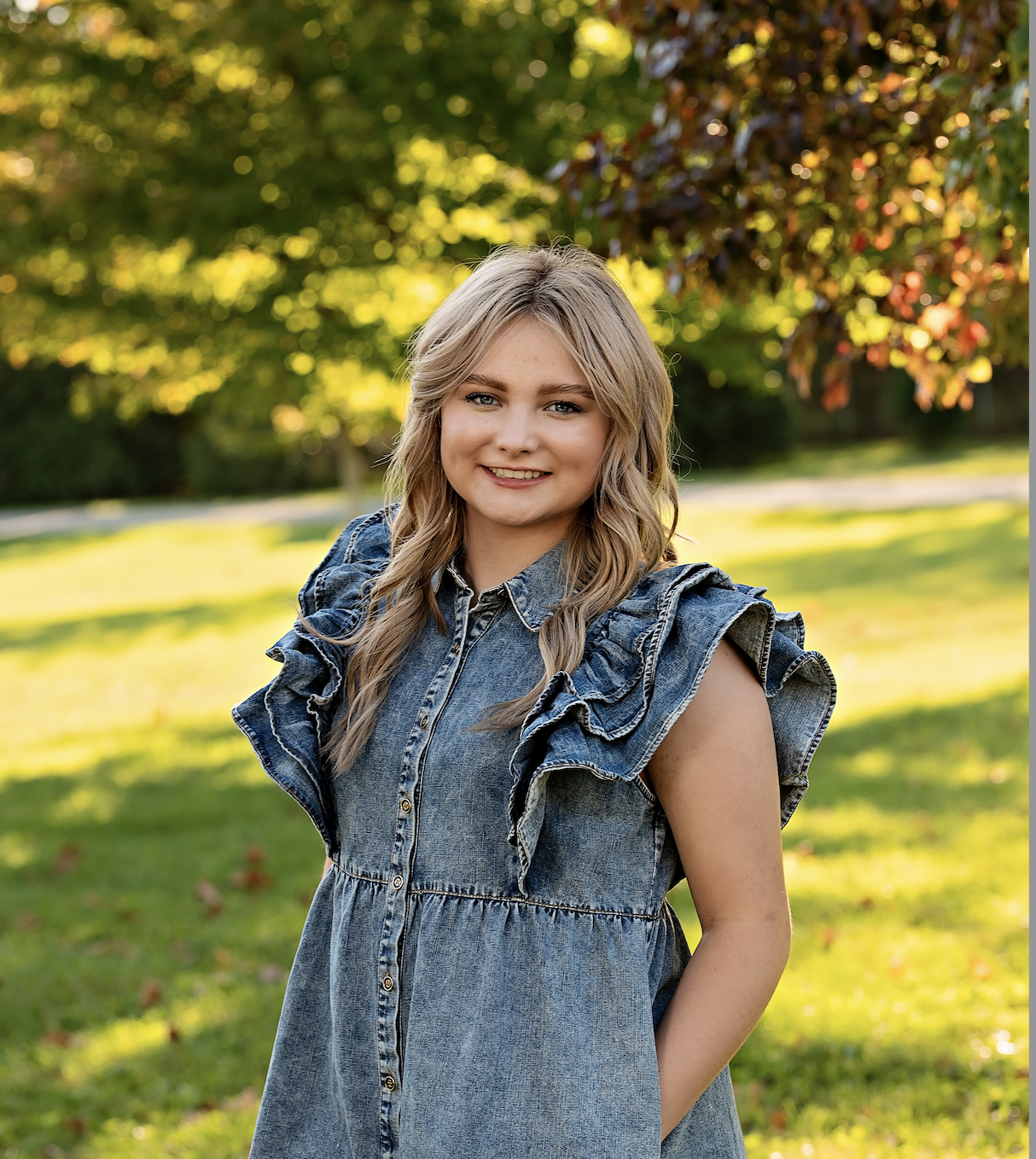 A young woman with blonde wavy hair smiling outdoors during daytime, wearing a denim dress with ruffled sleeves, in a park with green grass and trees with yellow and orange leaves.