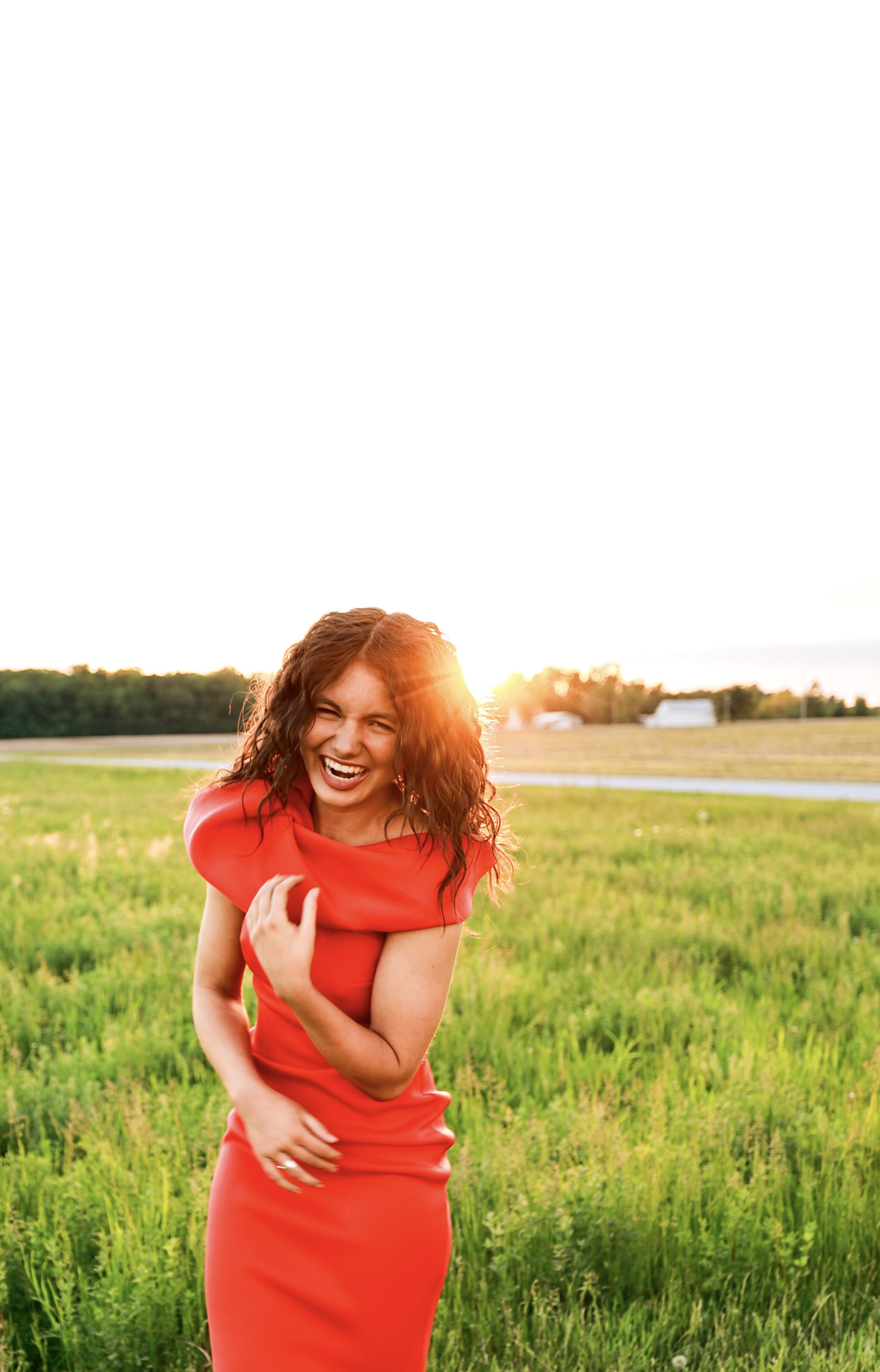 A woman in a bright red dress laughing and enjoying herself outdoors during sunset, standing in a green field.