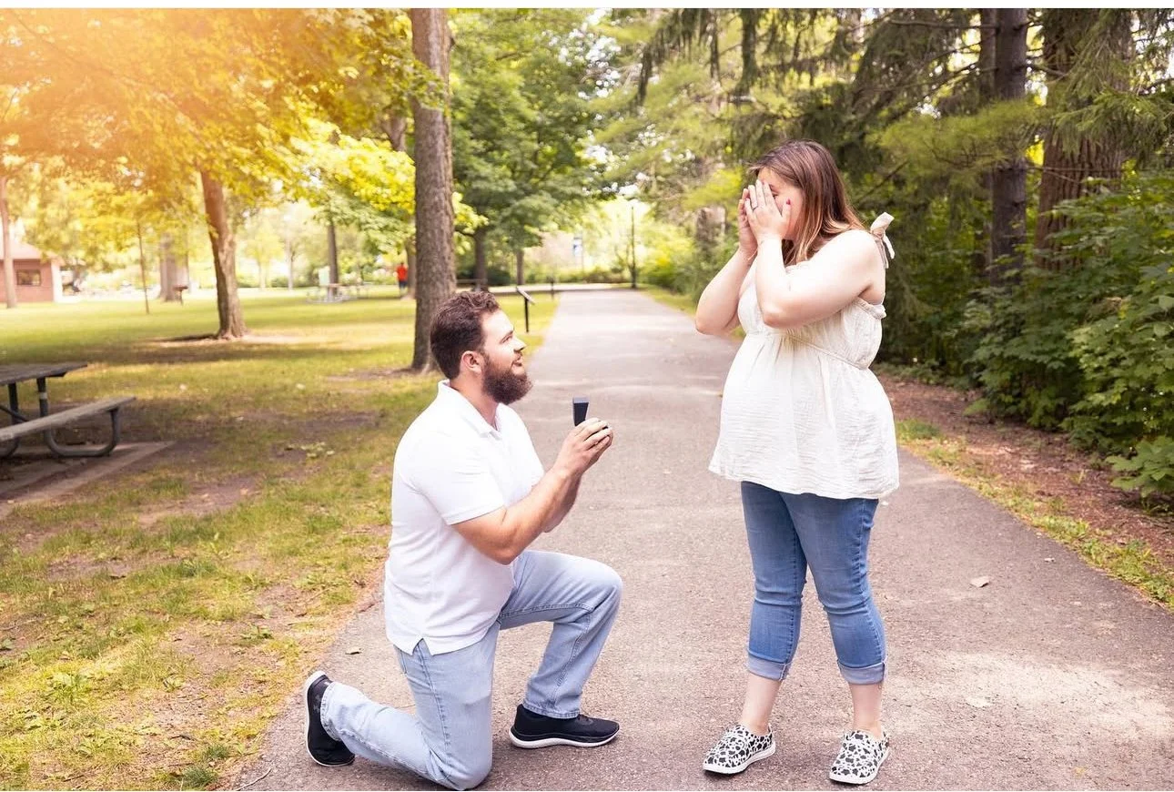 Man proposing on one knee with ring box to surprised woman on walking path in park.