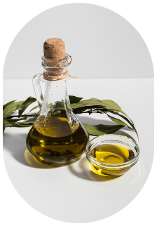A glass jar with a cork stopper filled with olive oil, a small glass bowl with more olive oil, and green leaves on a white surface.