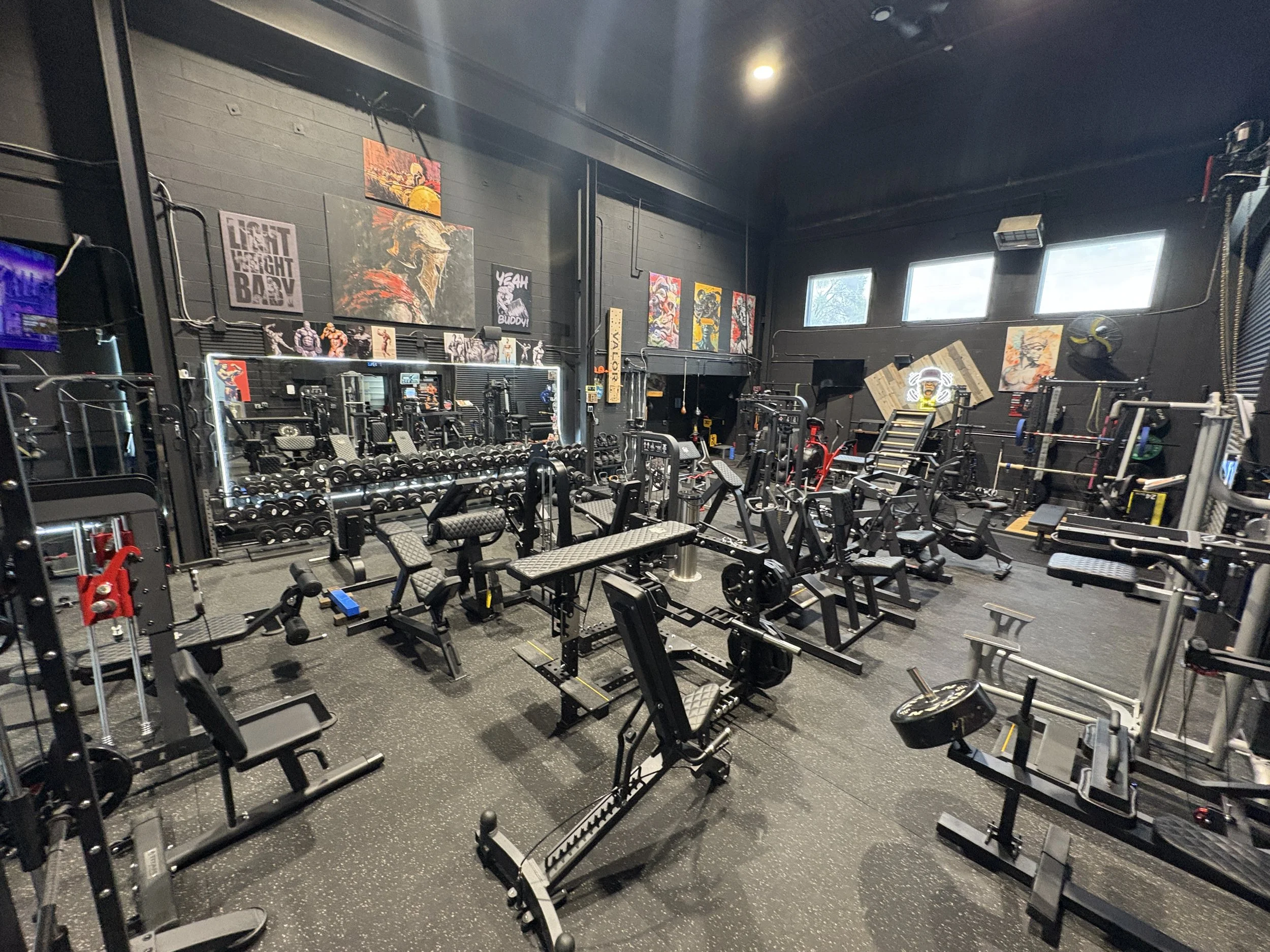 Interior of a gym with various workout equipment including benches, dumbbells, weight machines, and a mirror with posters on the black wall.