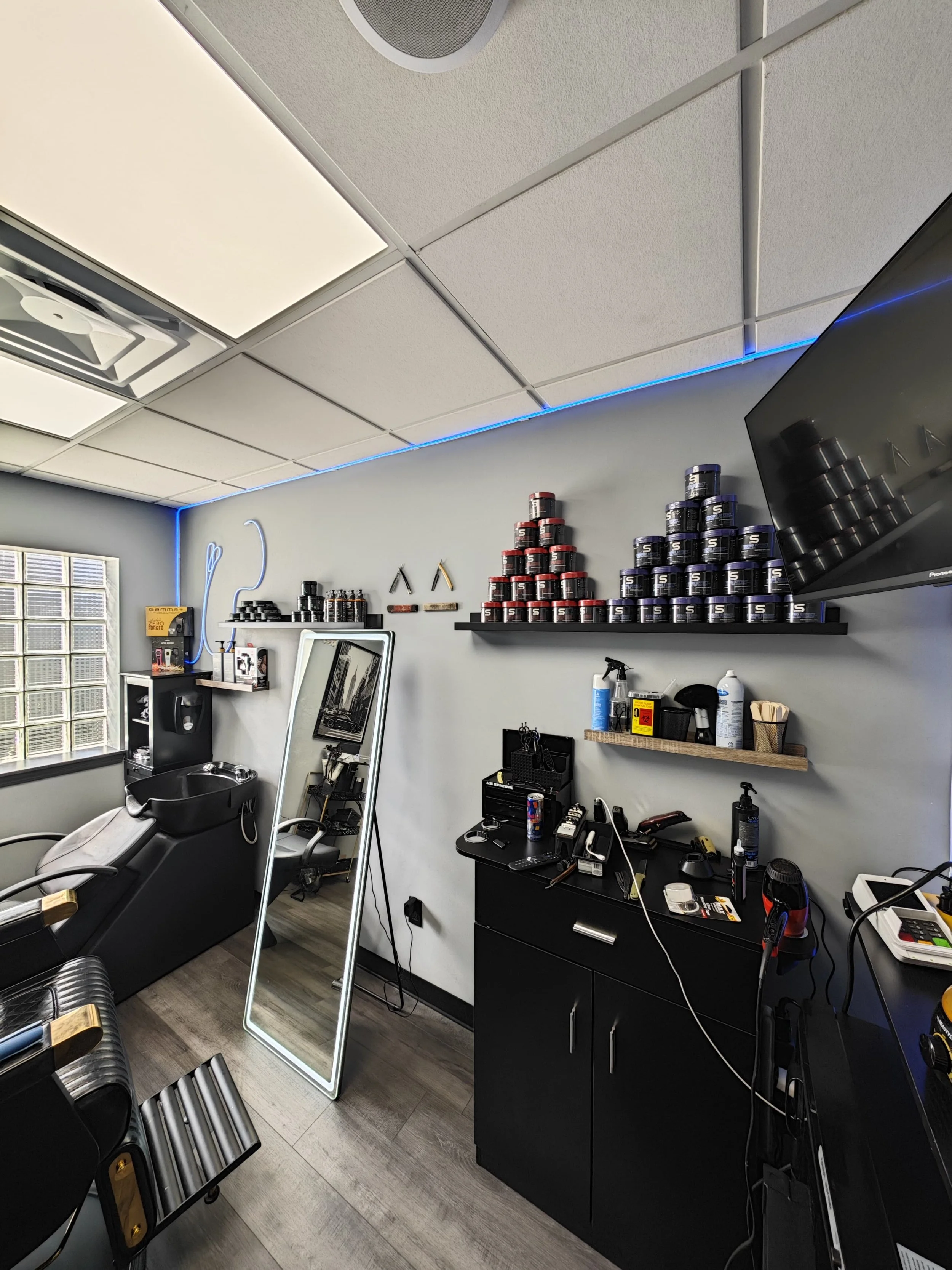 Interior of a barber shop or salon with a reclining barber chair, a large mirror, shelves of hair products, and various styling tools and supplies on counters and walls.