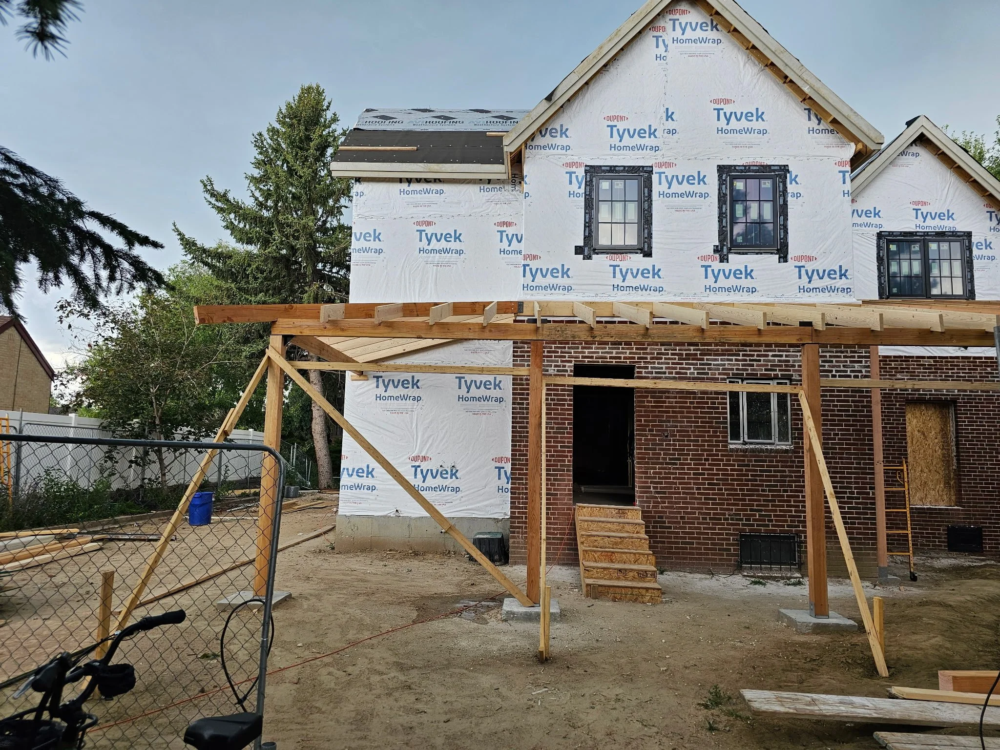 Under construction two-story brick house with wooden scaffolding and framing, construction materials, and a chain-link fence in the foreground.