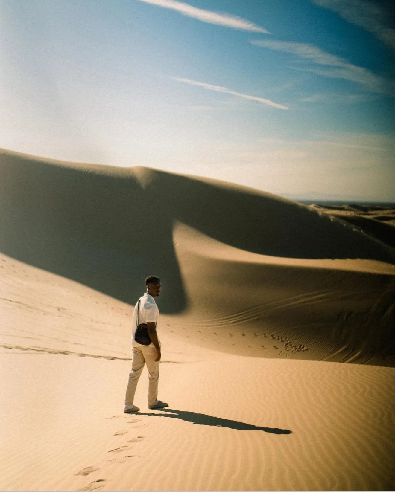 A person standing in a desert with sand dunes and a blue sky with wispy clouds in the background.