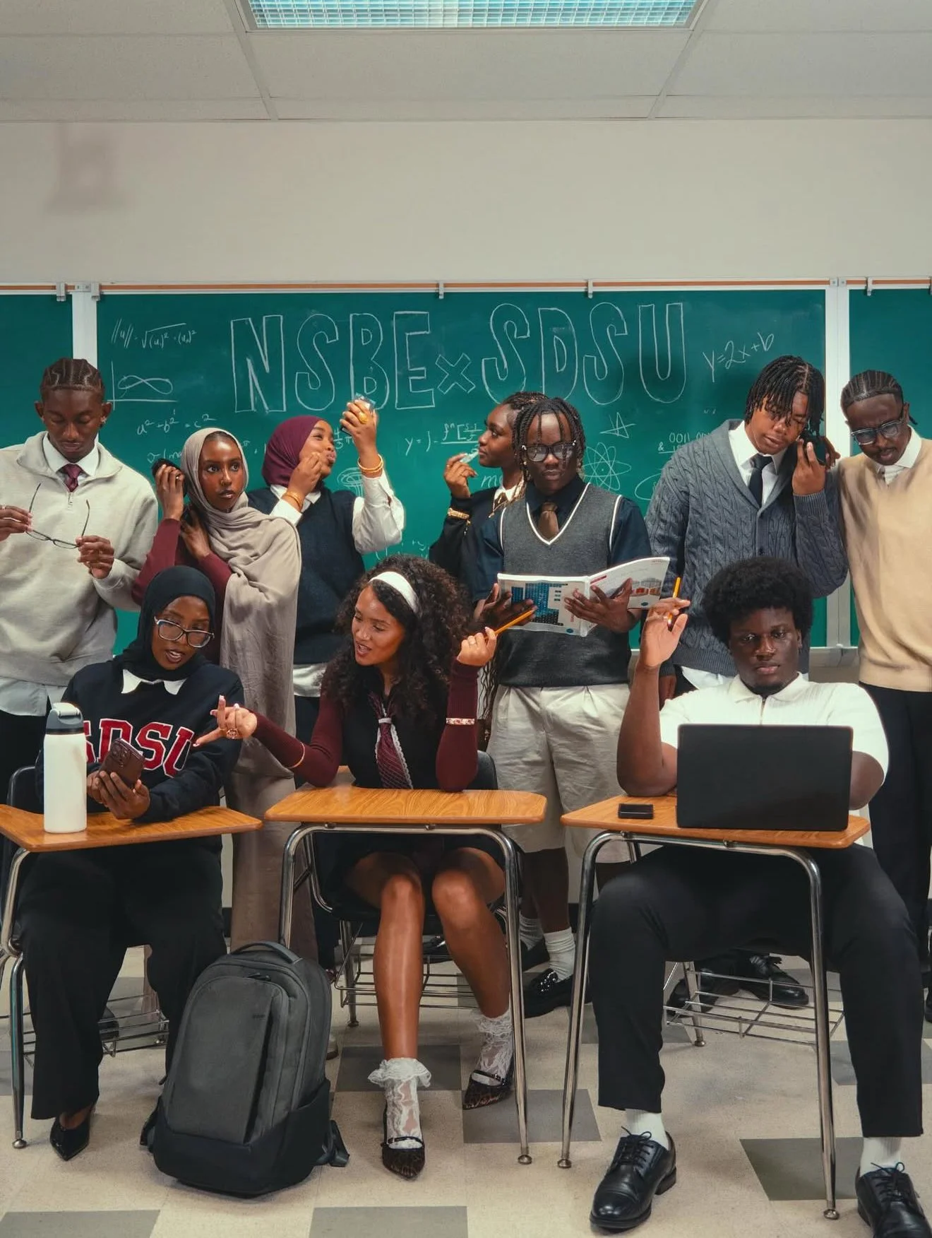 Classroom scene with two students seated at desks in the foreground and eight students standing behind them in front of a green chalkboard, which has various mathematical equations and symbols, and the text 'NSBE x SDSU' written on it.