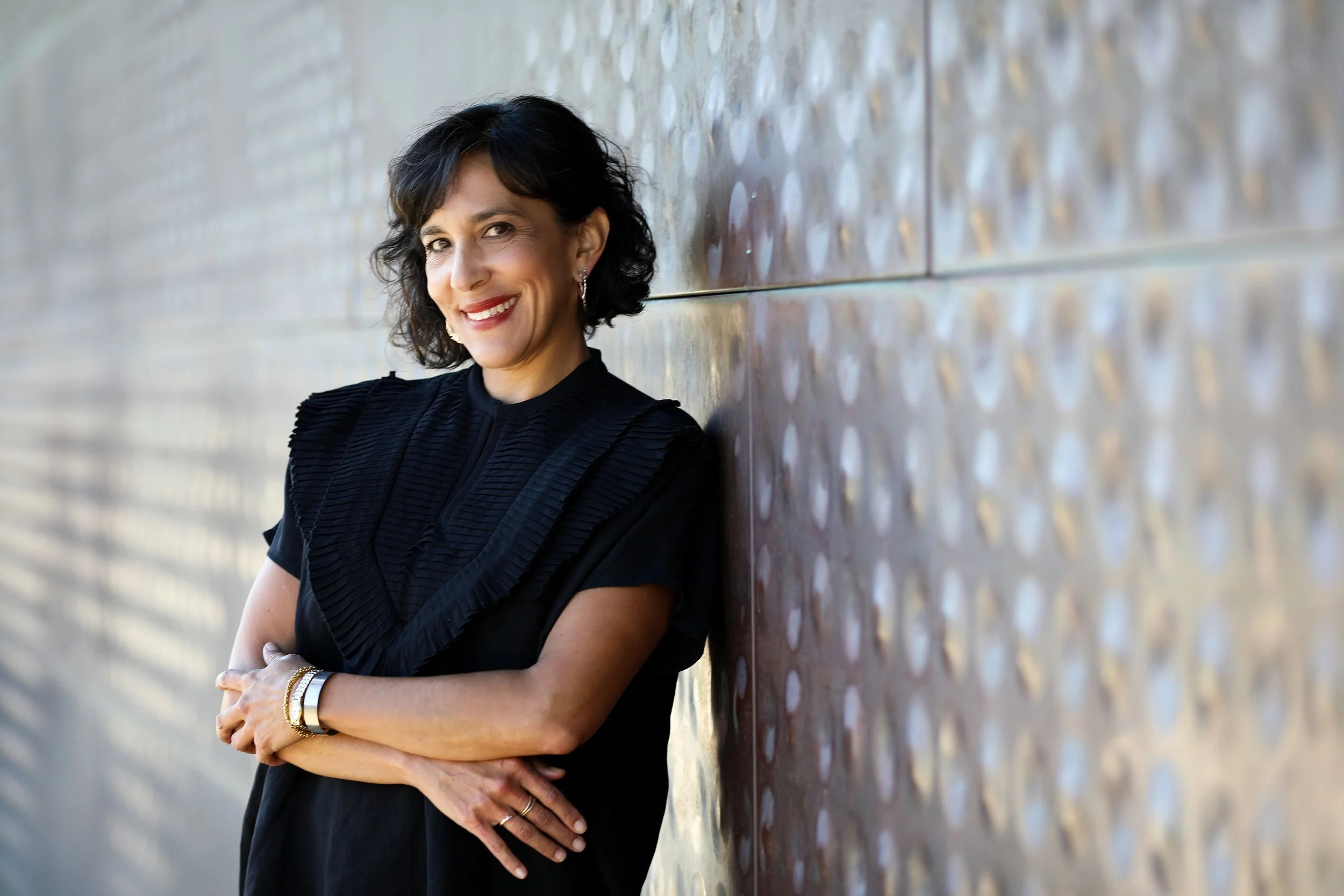 A woman with dark, wavy hair, smiling and wearing a black top, standing against a textured metallic wall.