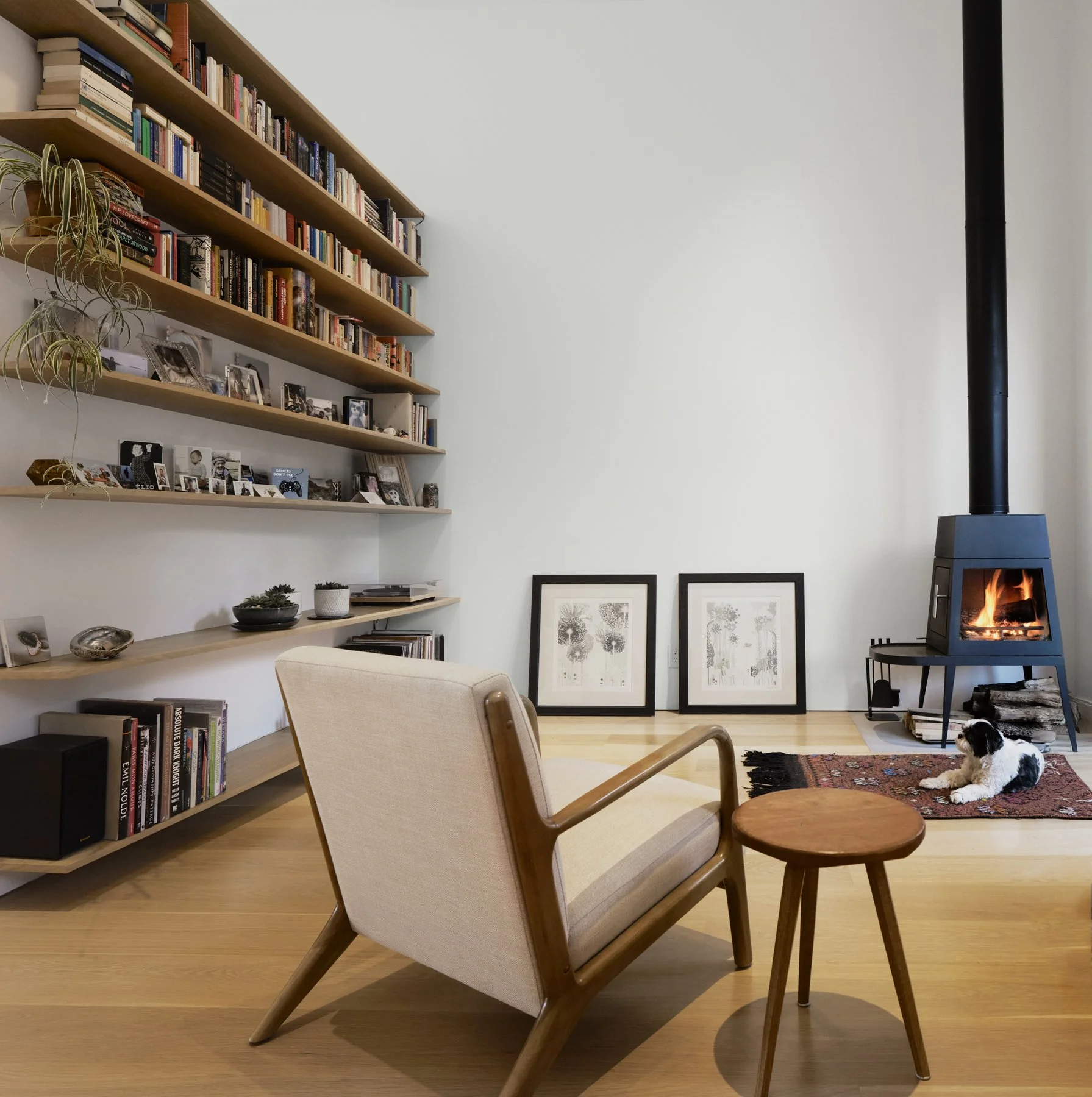 Living room with wooden floors, a beige armchair, and a small wooden stool, a fireplace with a fire burning, two framed pictures on the floor, a tall bookshelf filled with books and framed photos, plants, and decorative objects, and a black wood stove with logs underneath.