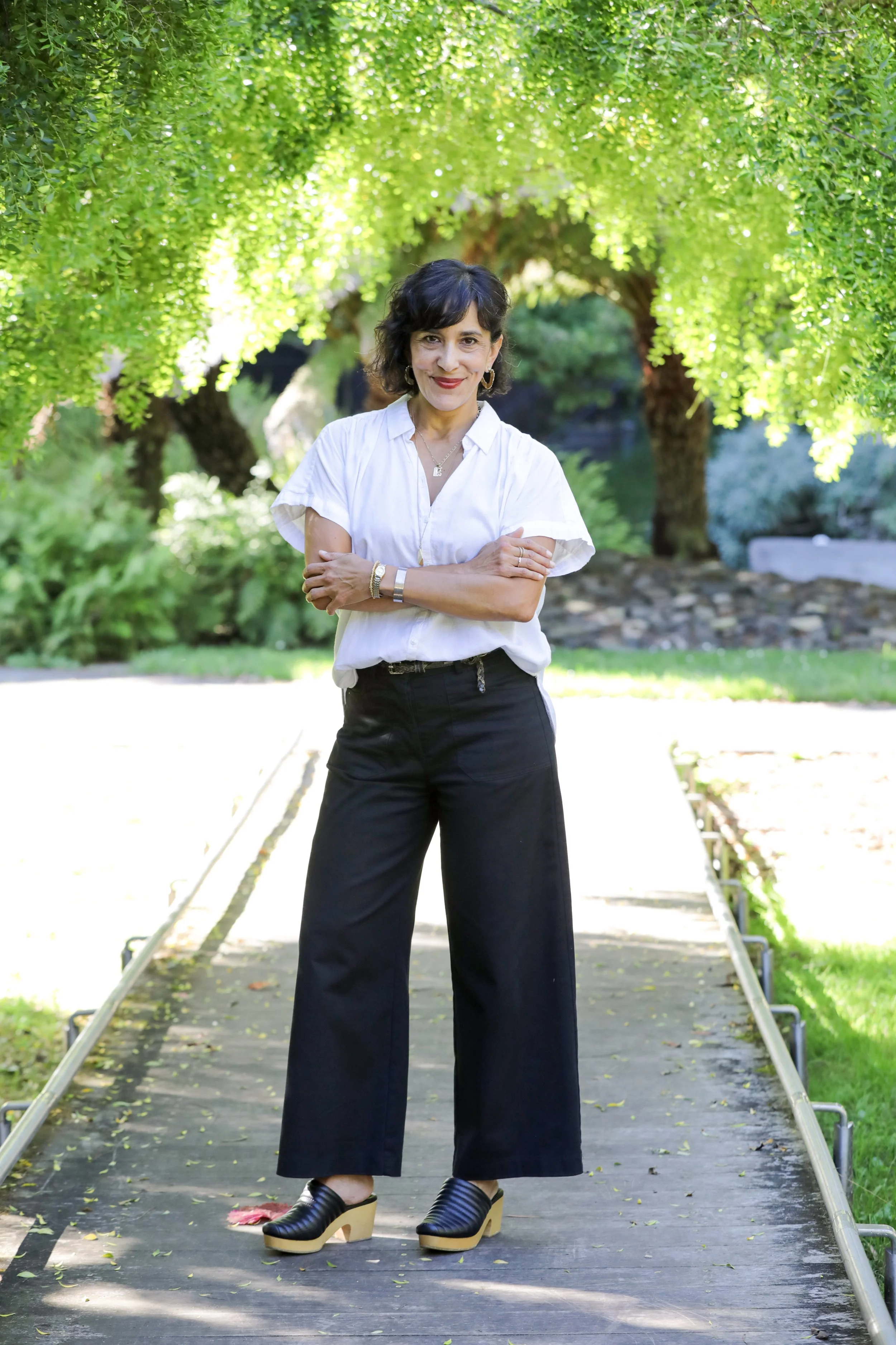 Woman standing on a small bridge in a lush green garden, smiling with arms crossed, wearing a white blouse, black wide-leg pants, and black heeled shoes.