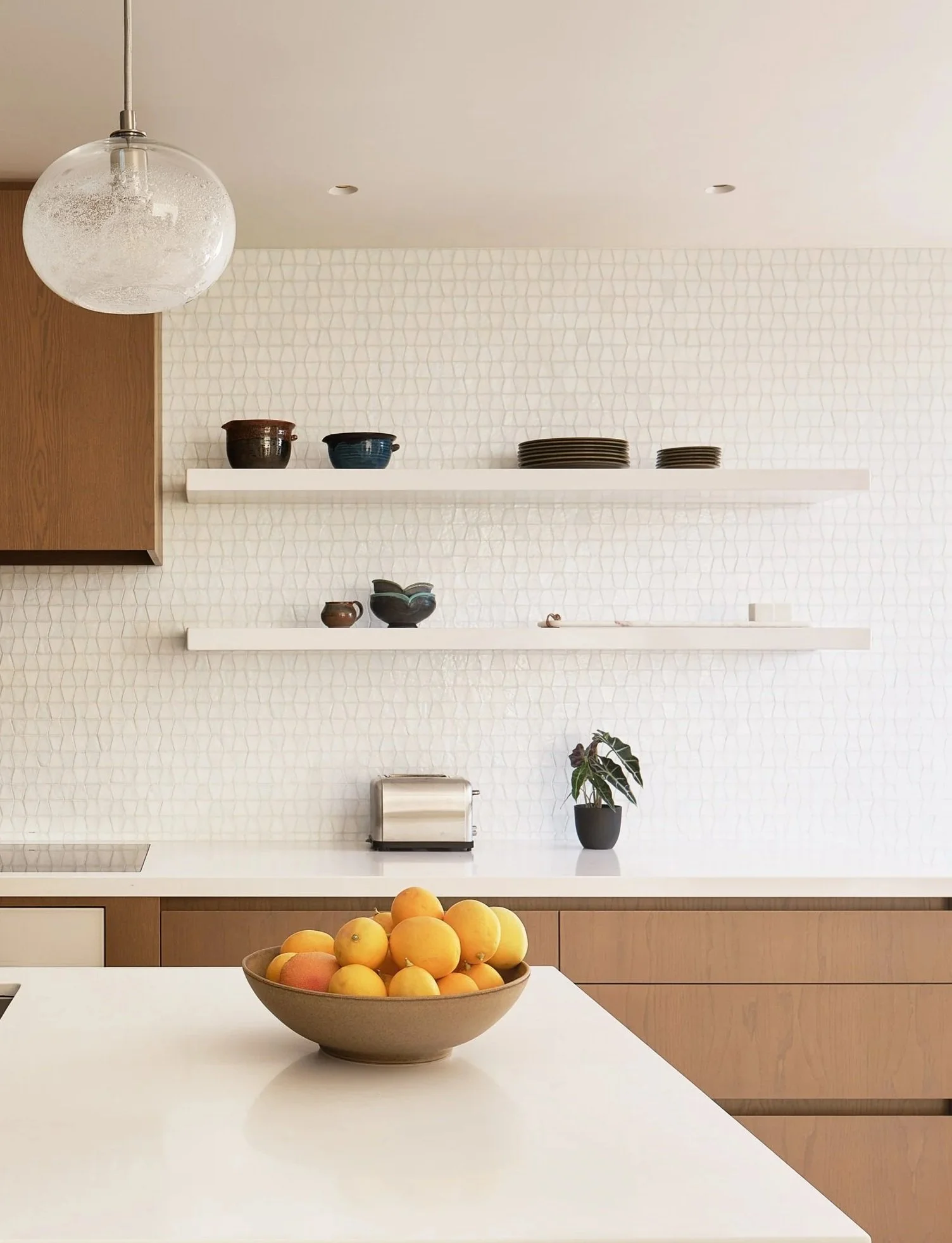 Kitchen with white countertops, wooden cabinets, white open shelves with dishes, a potted plant, a toaster, and a bowl of yellow and pink peaches on the counter.