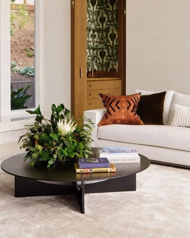 A living room with a white sofa, decorative pillows, a black coffee table with a floral arrangement, and books, with a window showing outdoor greenery in the background.