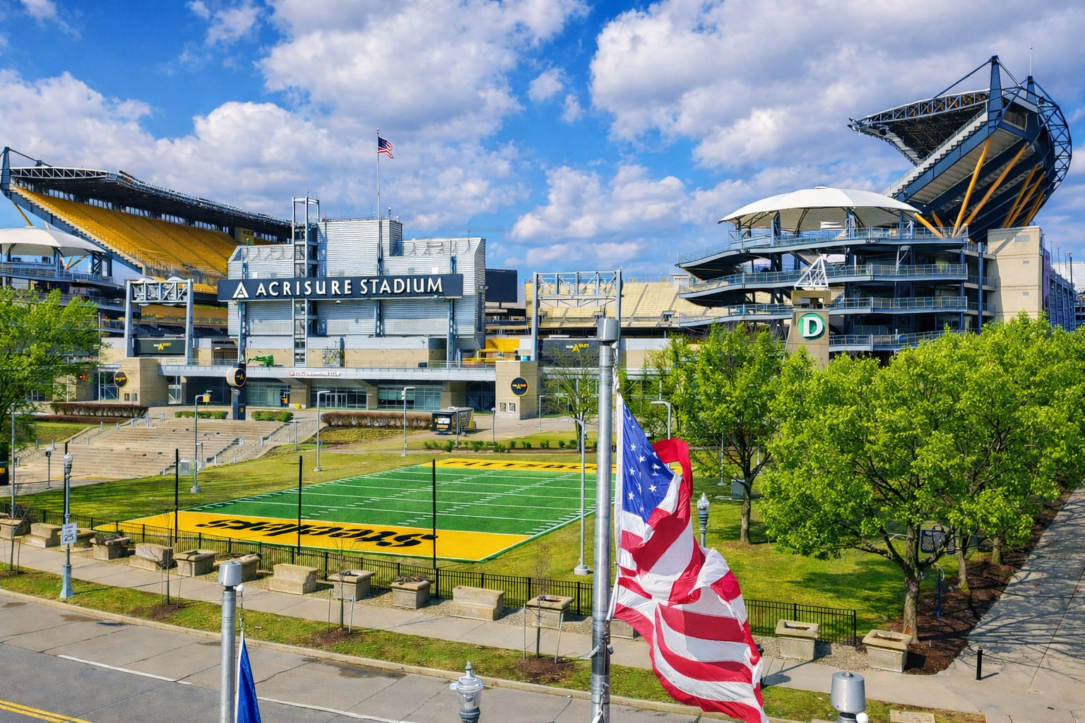 Acrisure Stadium with green trees, American flags, and a football field in front on a sunny day.