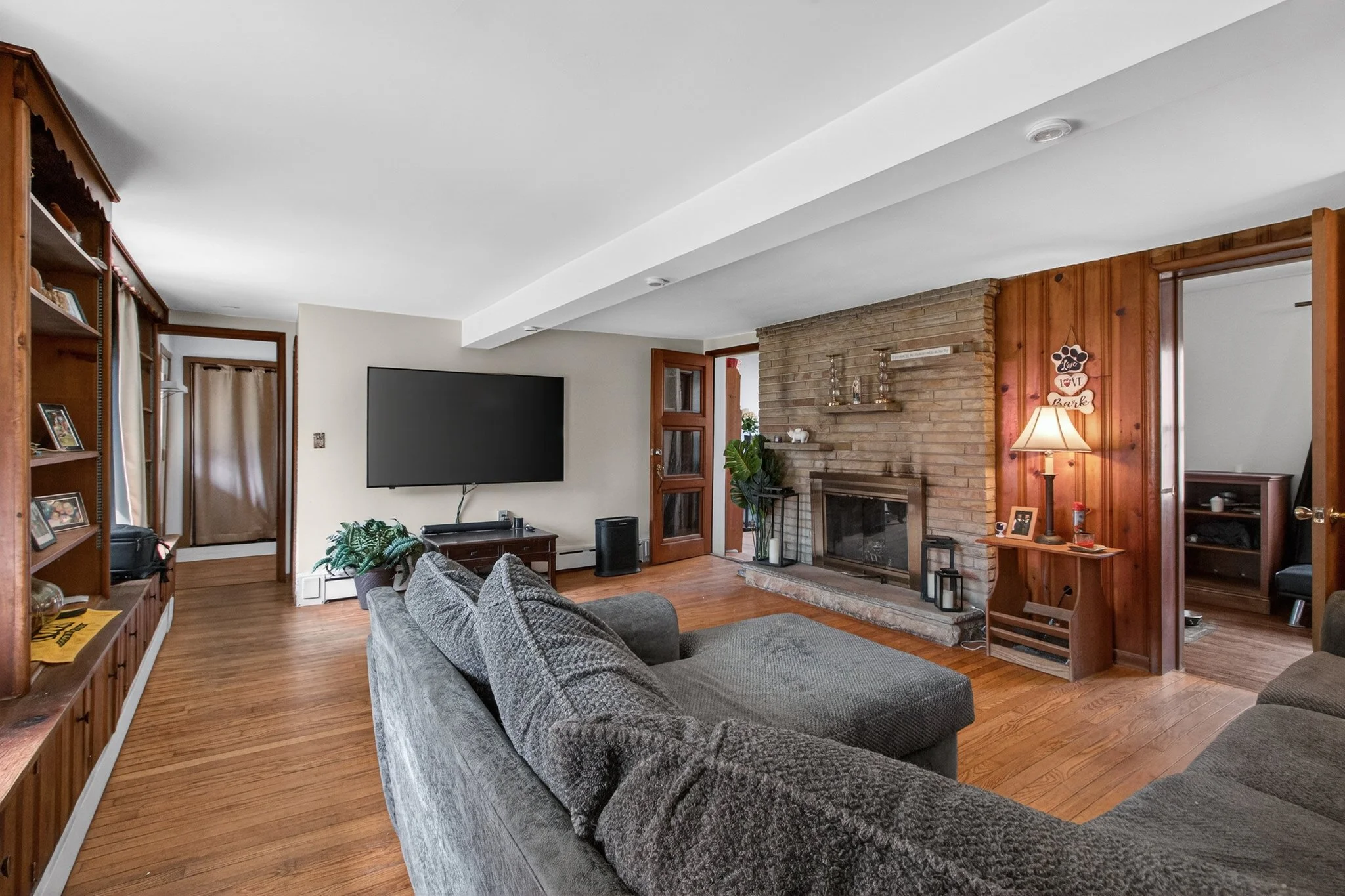 Living room with wooden floors, gray sectional sofa, brick fireplace, wall-mounted TV, and wooden shelves.