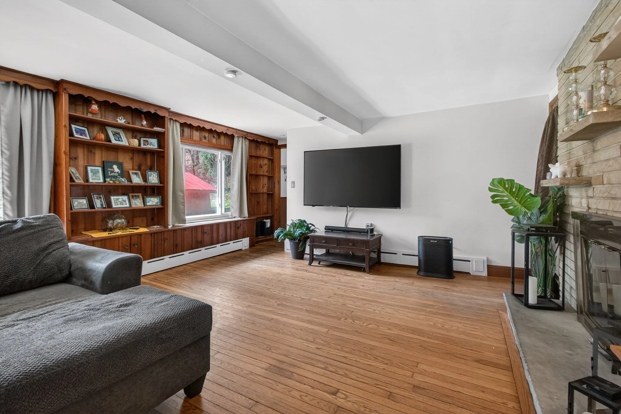 Living room with hardwood floors, gray sofa, wooden built-in shelves with family photos, a window with curtains, a large flat-screen TV on a stand, potted plants, and a brick fireplace with decorative items.