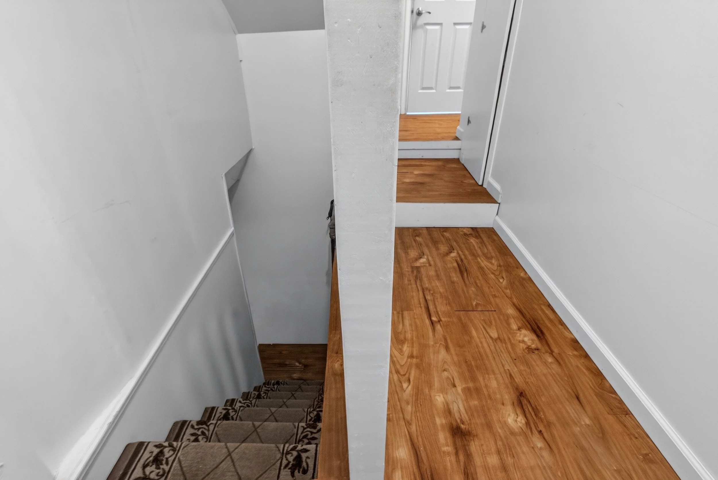 View of a staircase with carpeted steps on the left and a wooden-floored hallway with stairs on the right, separated by a white wall and a partial column. There is a white door at the top of the right staircase.
