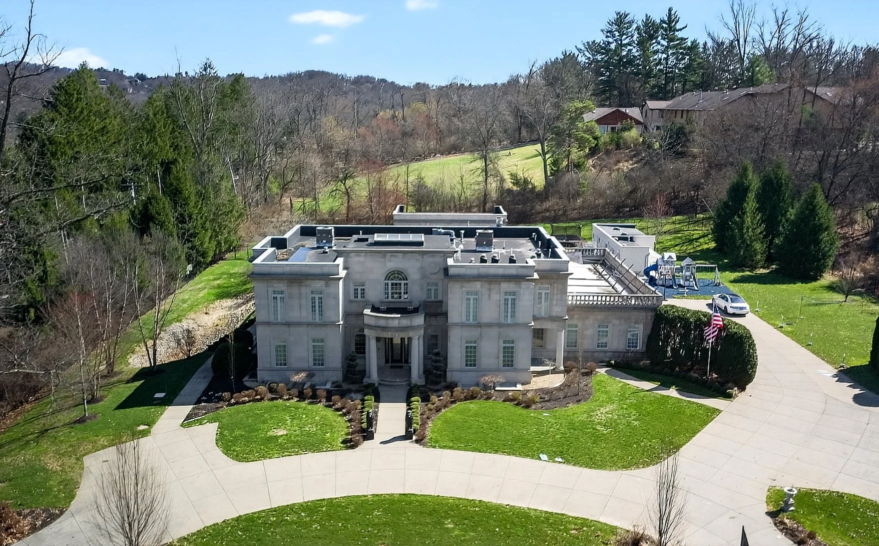 A large, white mansion with a grand entrance featuring columns, surrounded by well-maintained lawns, shrubs, and trees, with a curved driveway and a children's playground in the backyard.