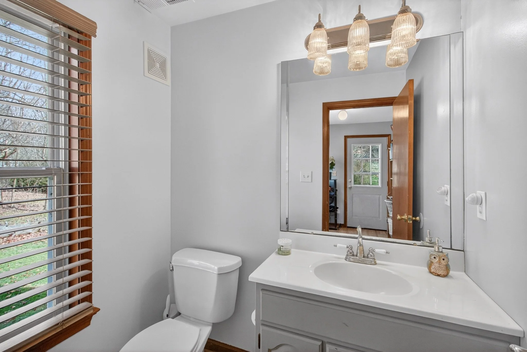 Bathroom with a white toilet, white sink, large mirror, and a window with wooden blinds, visible in a doorway.