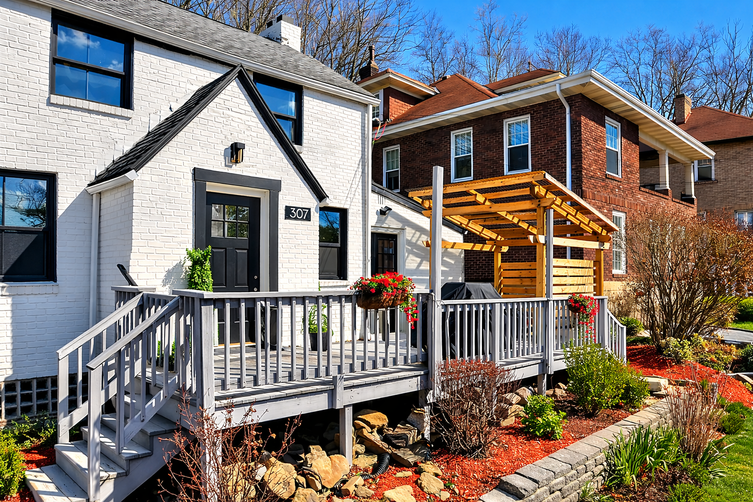 White brick house with black door and window frames, with a wooden deck and outdoor seating area, surrounded by landscaping and garden plants.