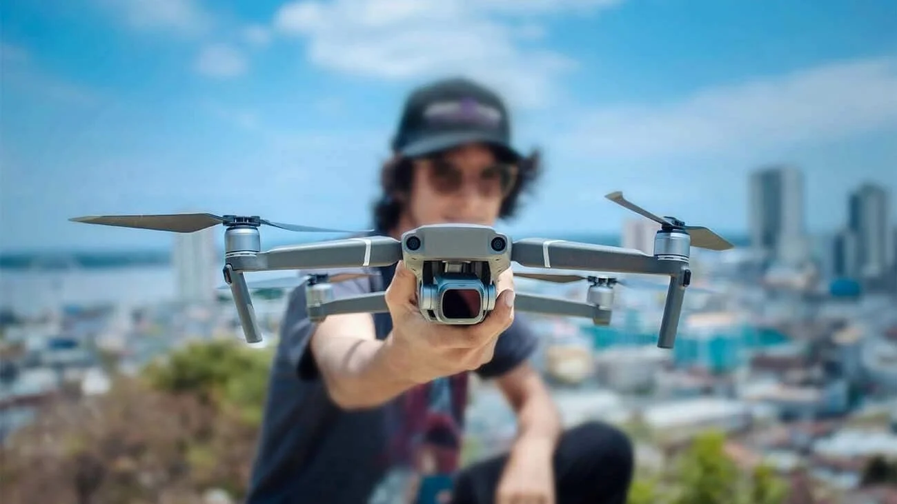 Person wearing sunglasses and a cap flying a drone with a cityscape in the background.