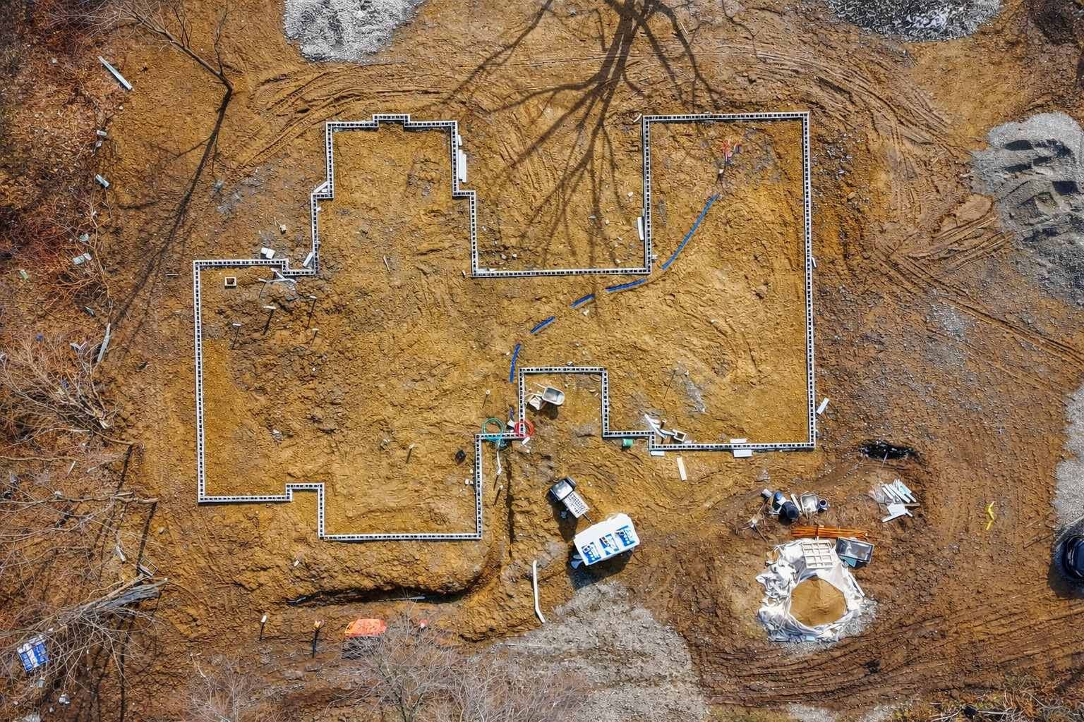 Aerial view of a house foundation under construction on a dirt lot. The foundation walls are outlined with metal framing, and there are construction tools, debris, and equipment scattered around.