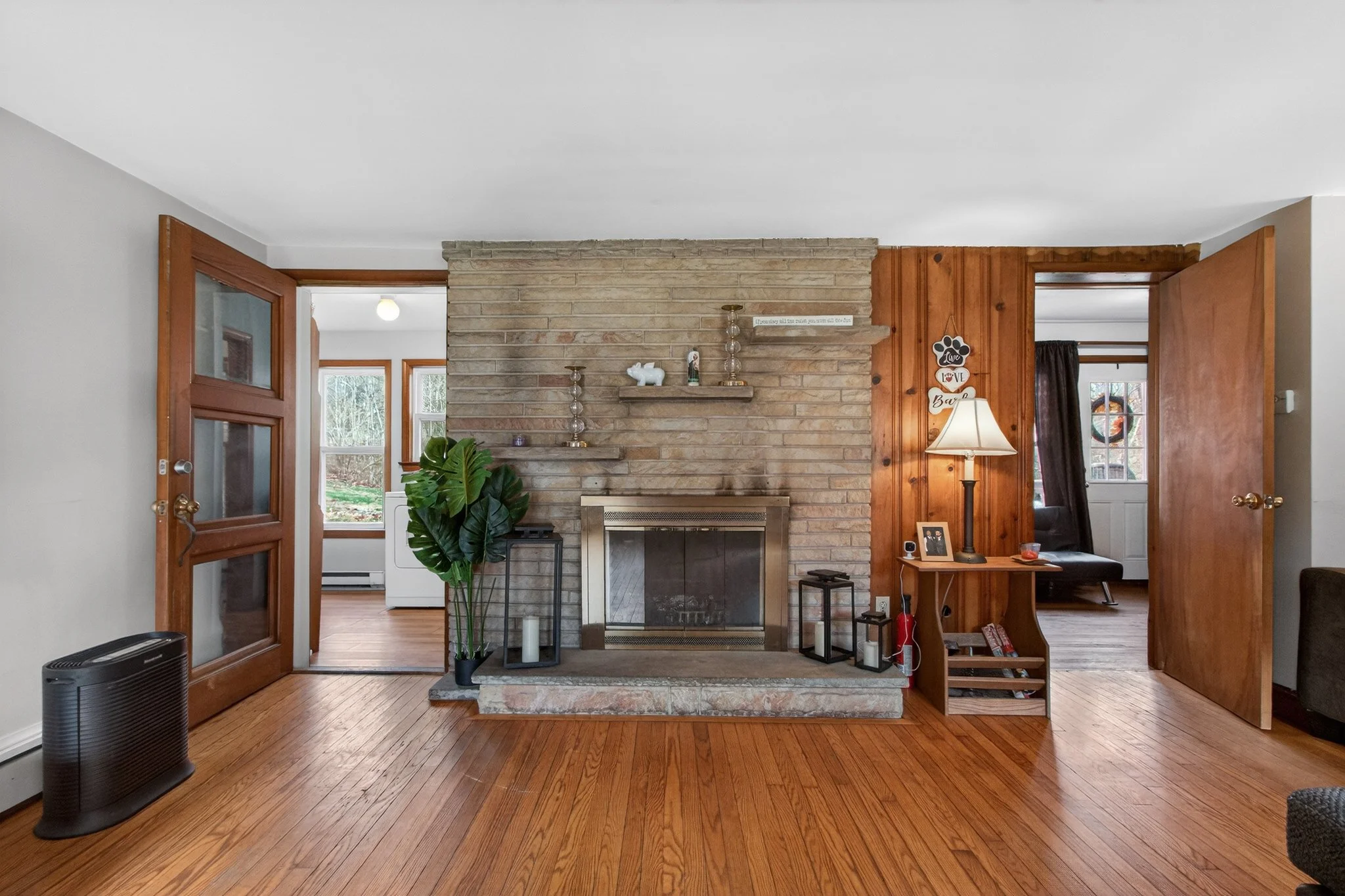 Living room with a brick fireplace, wooden floors, and two doorways leading to other rooms, decorated with a large green plant, candles, and various small decor items.
