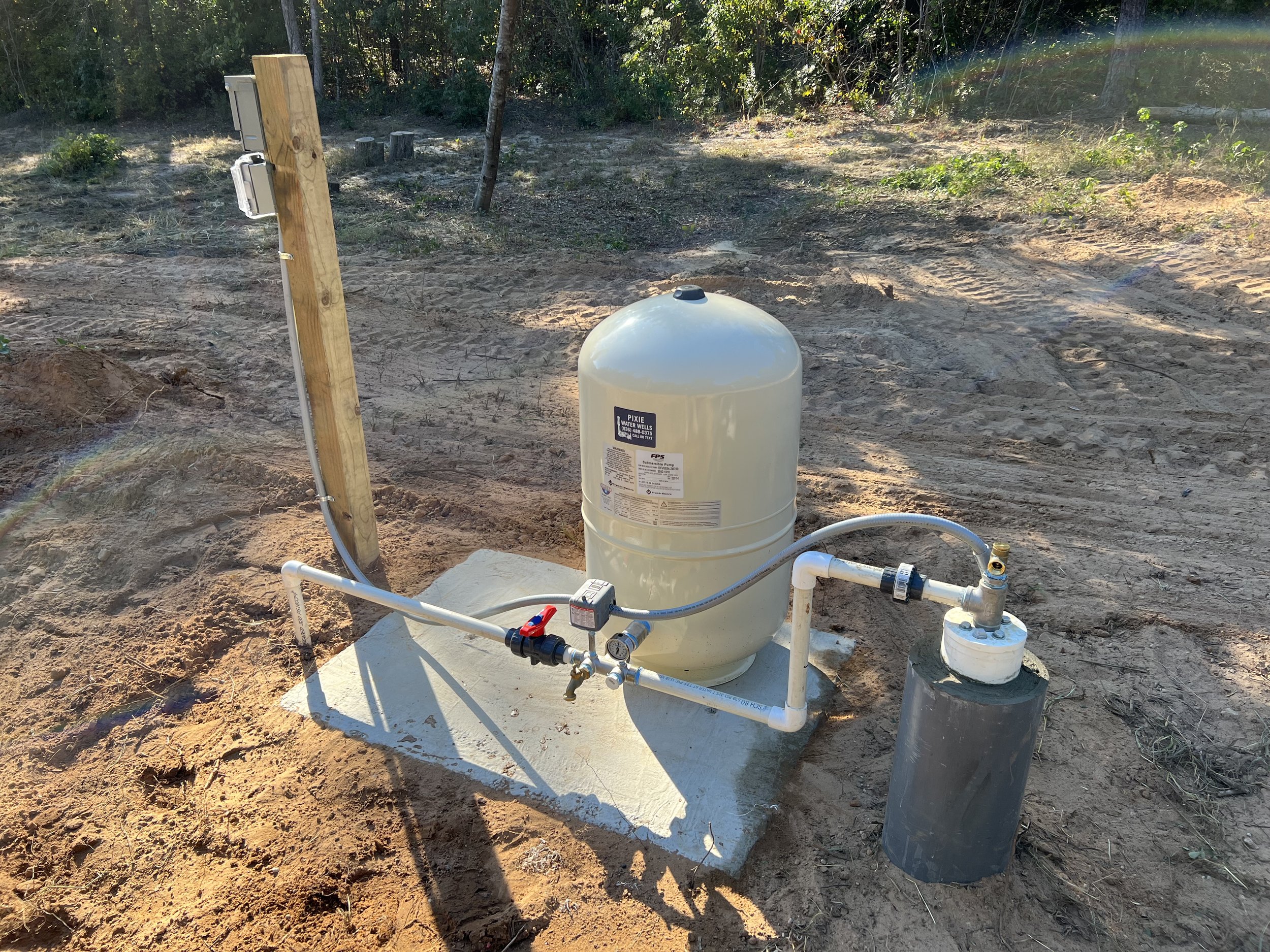 An outdoor water pressure tank system installed on a concrete pad with pipes, valves, and a gauge, surrounded by dirt and trees.