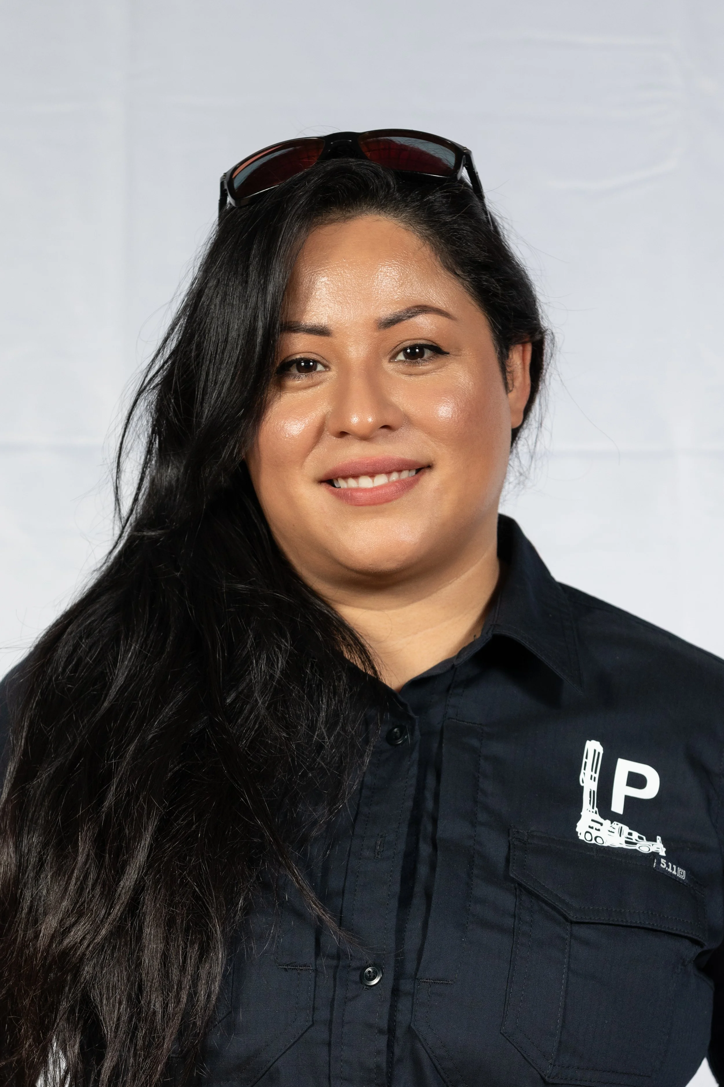 A woman with long dark hair wearing sunglasses on her head and a dark uniform shirt with a white logo that says 'P' and depicts a drilling rig, standing against a light background.