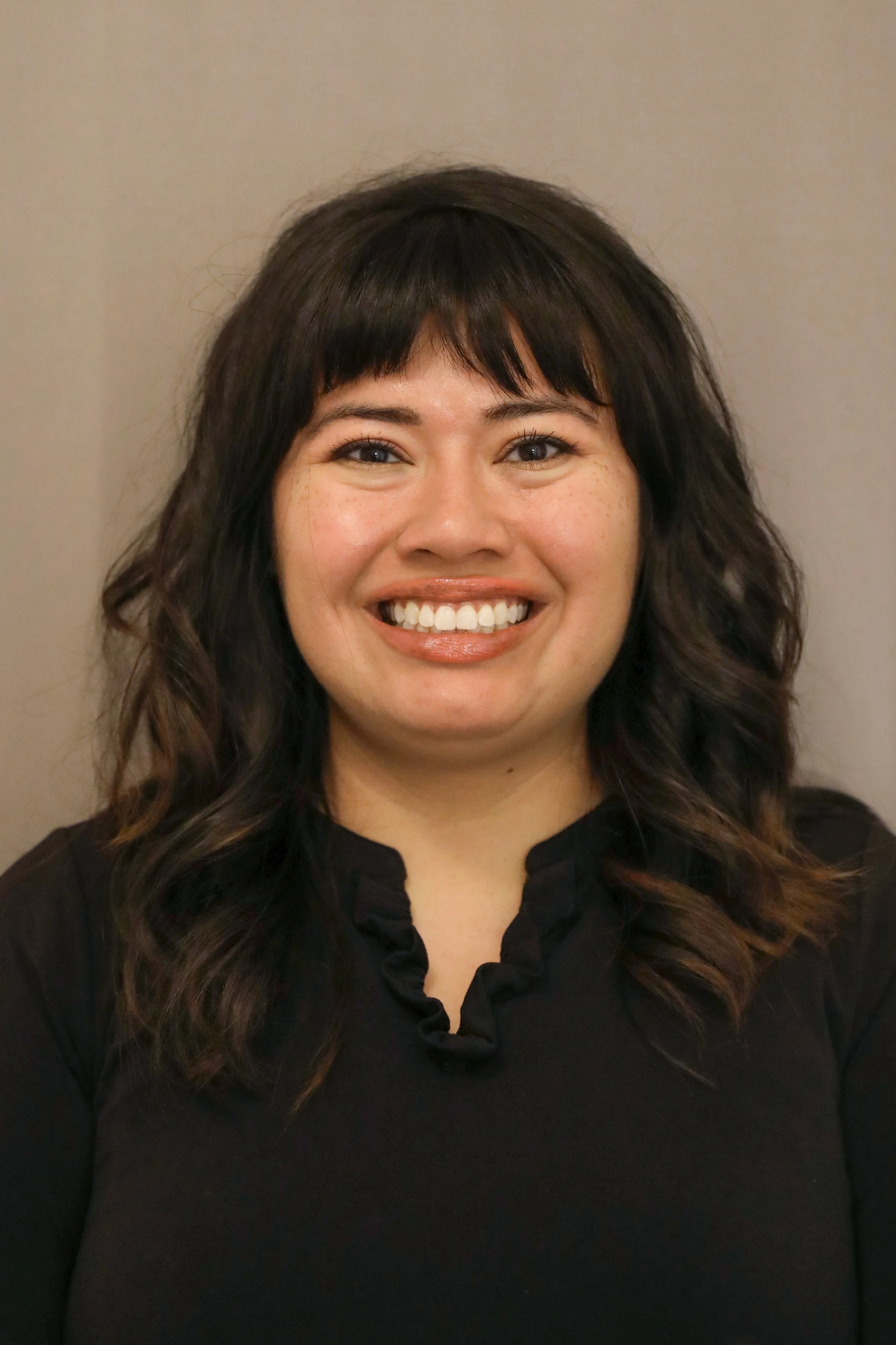 A woman with dark wavy hair, smiling and showing her teeth, wearing a black top with ruffled collar standing against a plain background.