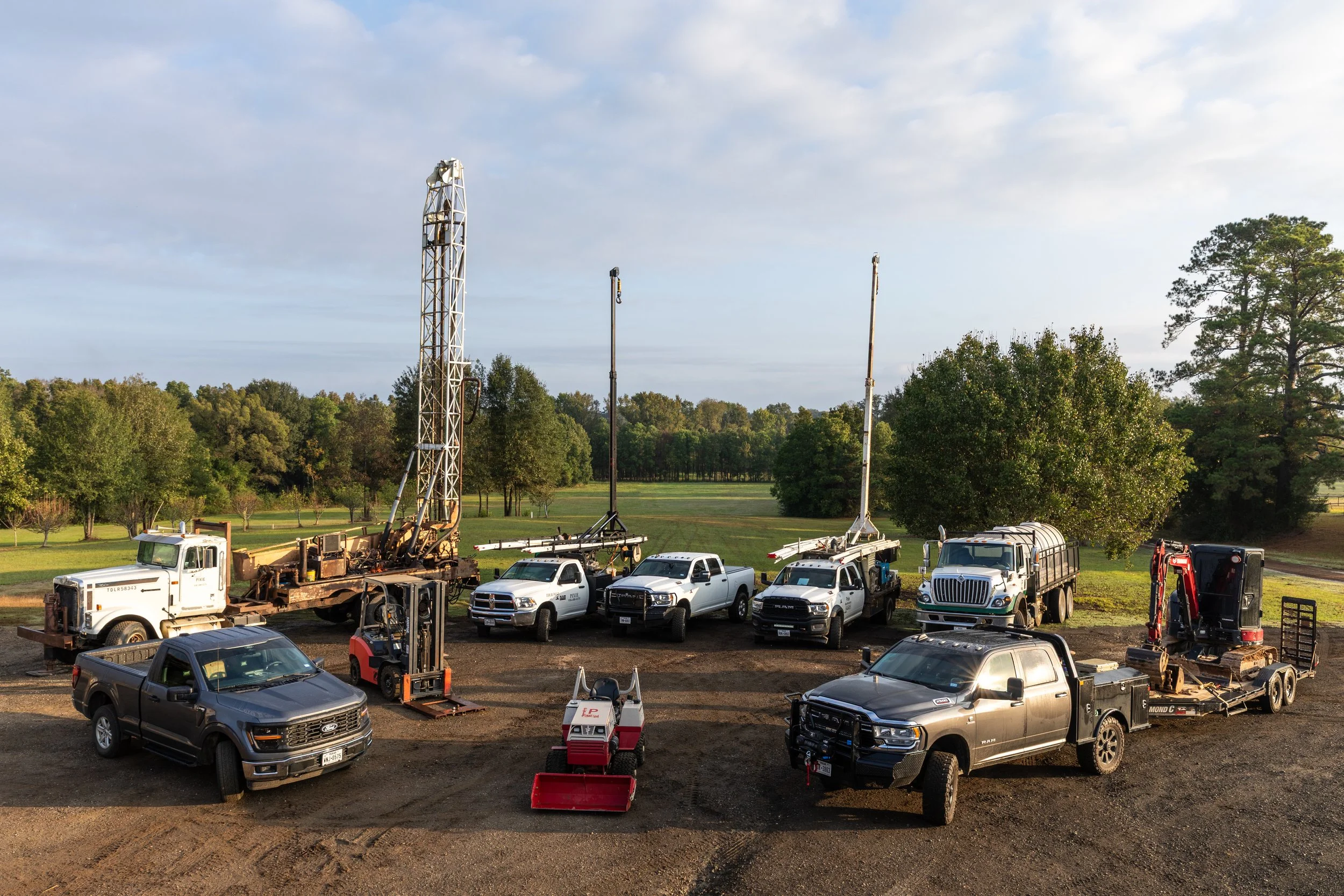 A collection of construction and utility vehicles, including trucks, excavators, and forklifts, parked on a dirt lot with a grassy field and trees in the background during daytime.