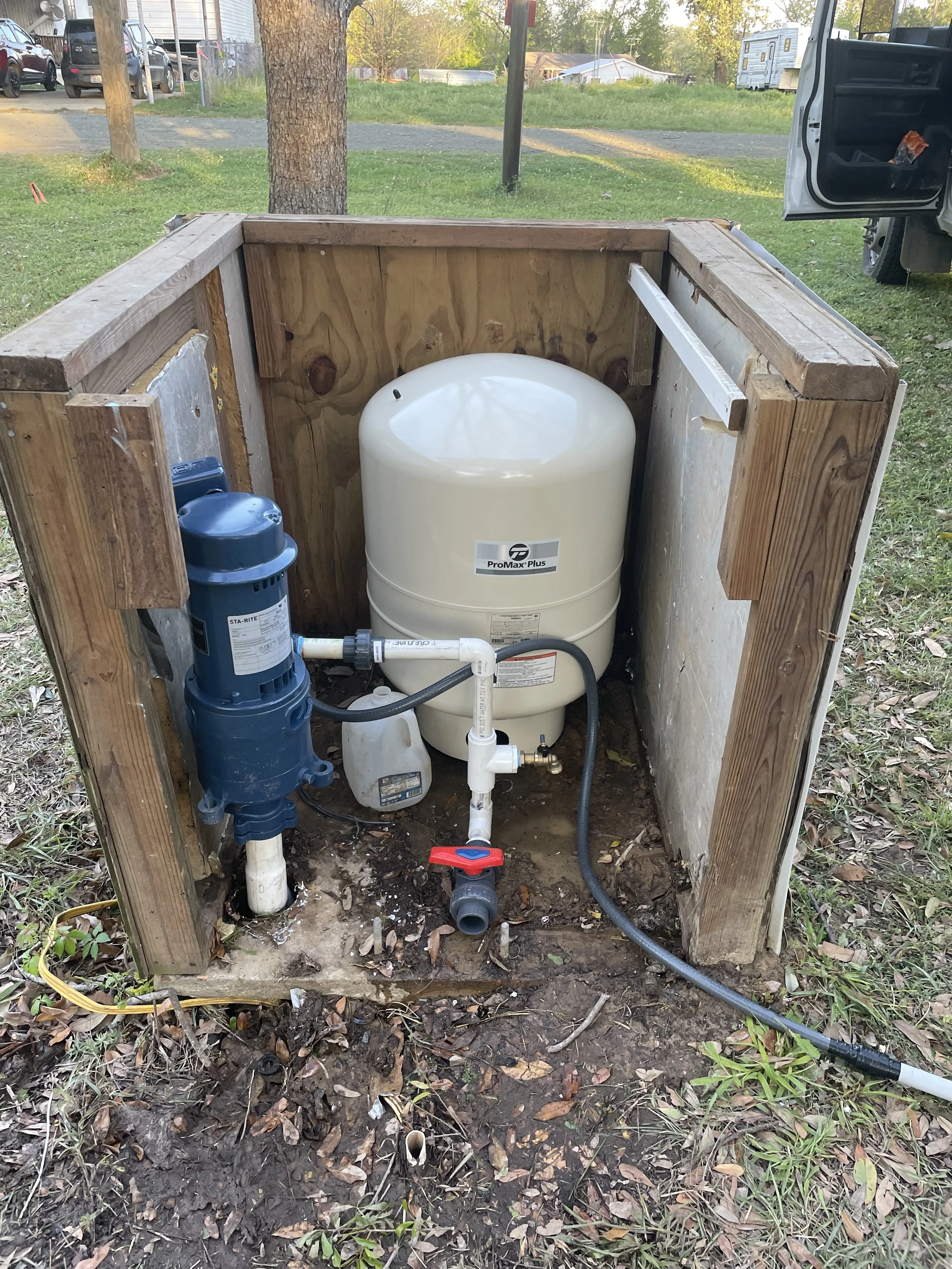 An outdoor water pressure tank system installed inside a wooden enclosure with pipes and a blue pump.