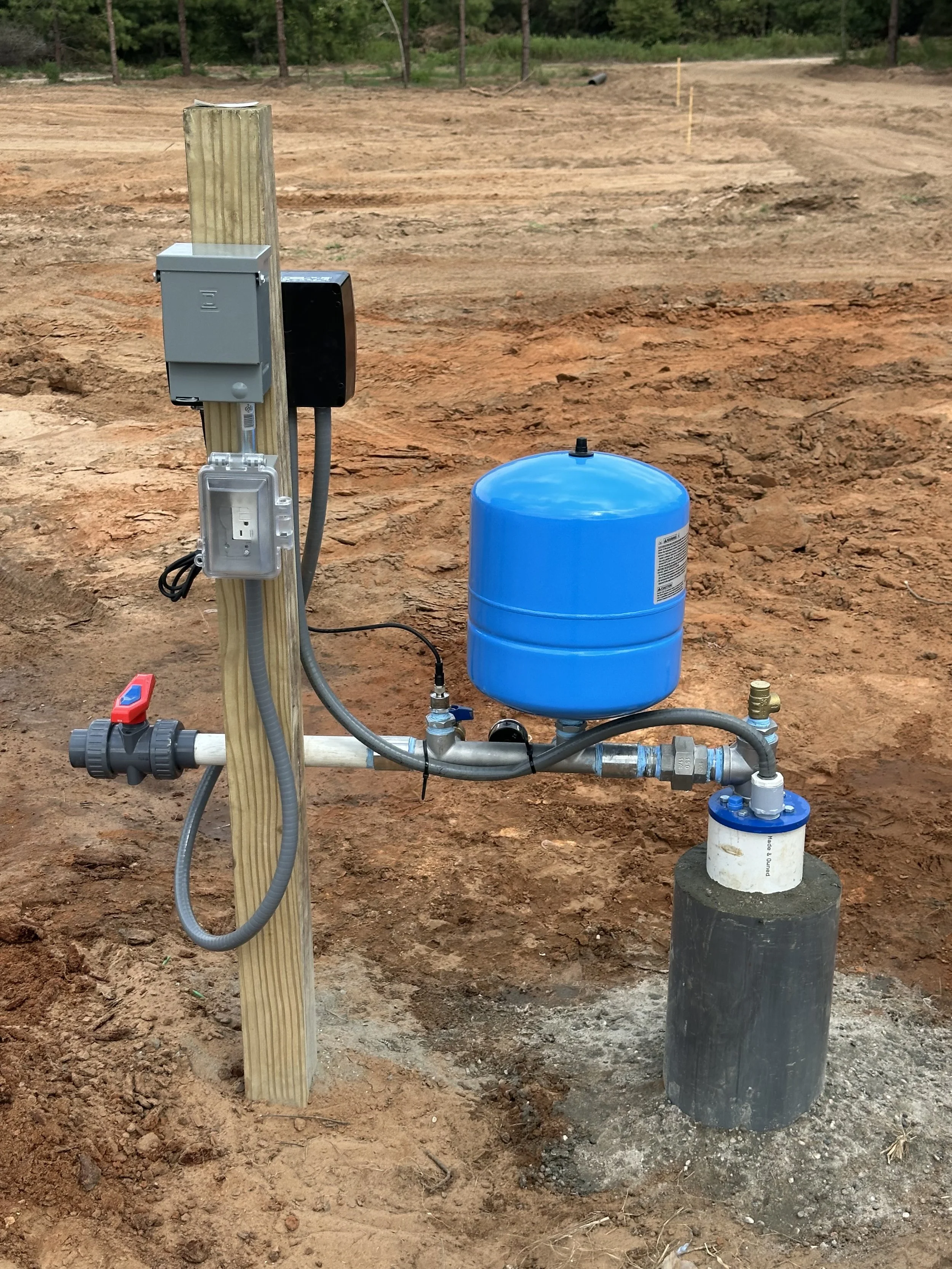A water pressure tank system installed outdoors on a dirt surface, with electrical components mounted on a wooden post.