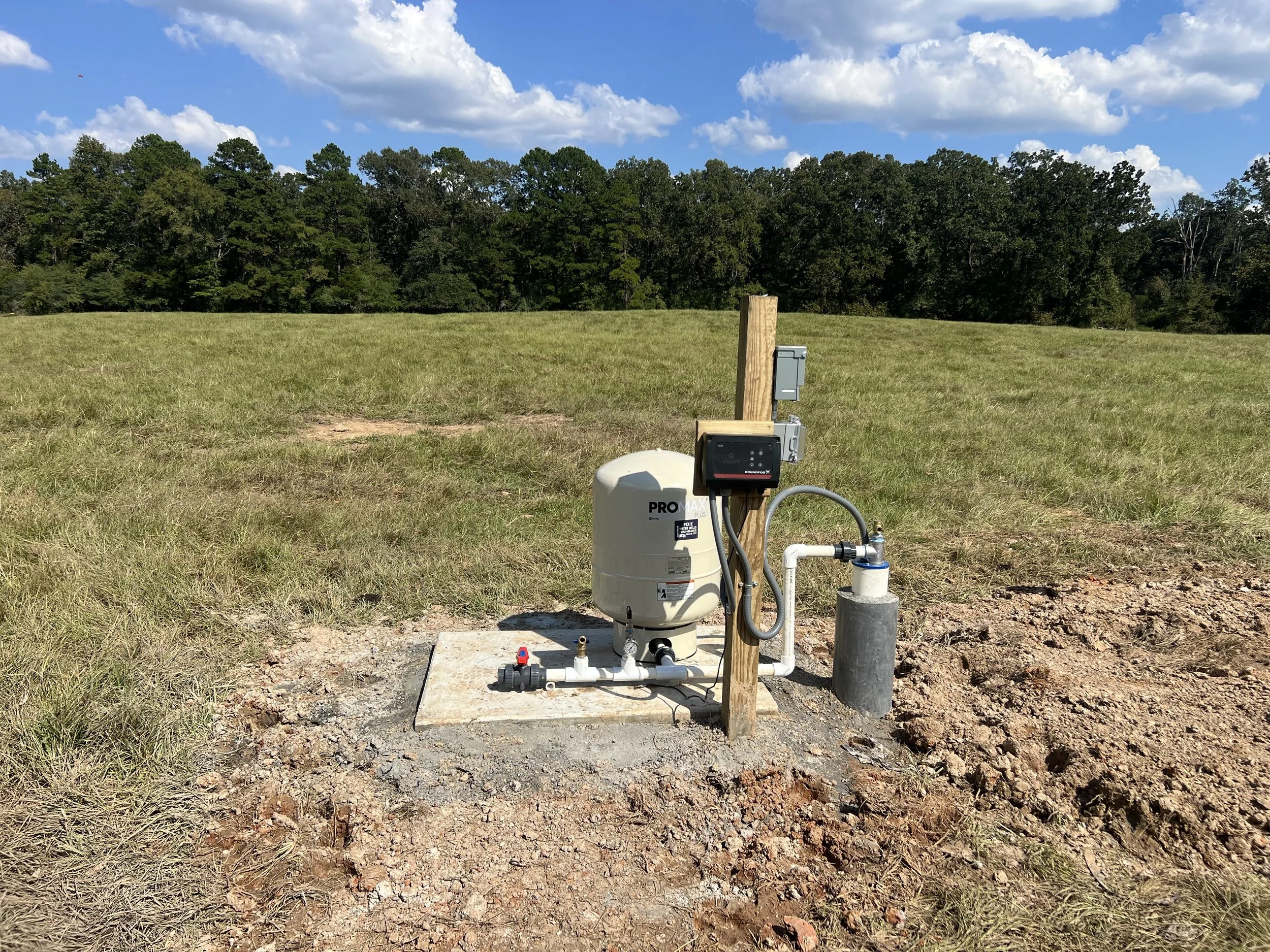 An outdoor water well pump system on a concrete pad in a grassy field with trees and a partly cloudy sky in the background.