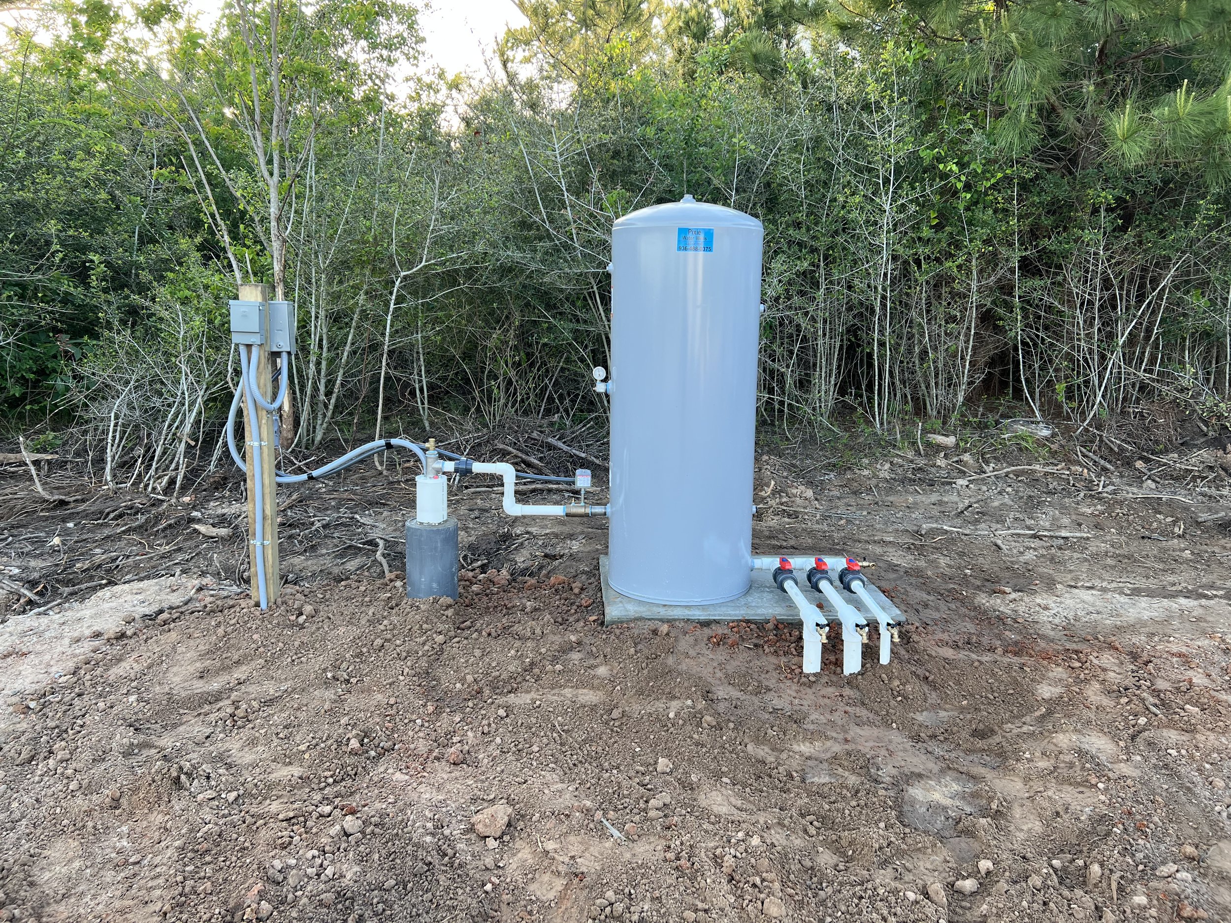 A large gray hydro-pneumatic tank installed outdoors on a concrete pad, connected to electrical box and plumbing pipes, set against a backdrop of trees and bushes.