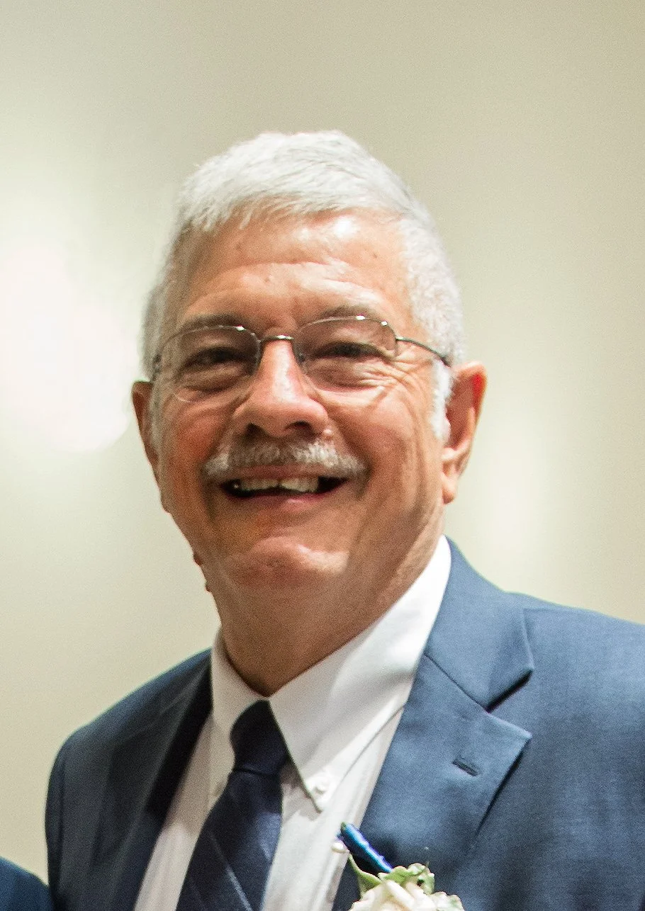 Smiling older man with glasses, wearing a suit and tie, in front of plain wall.