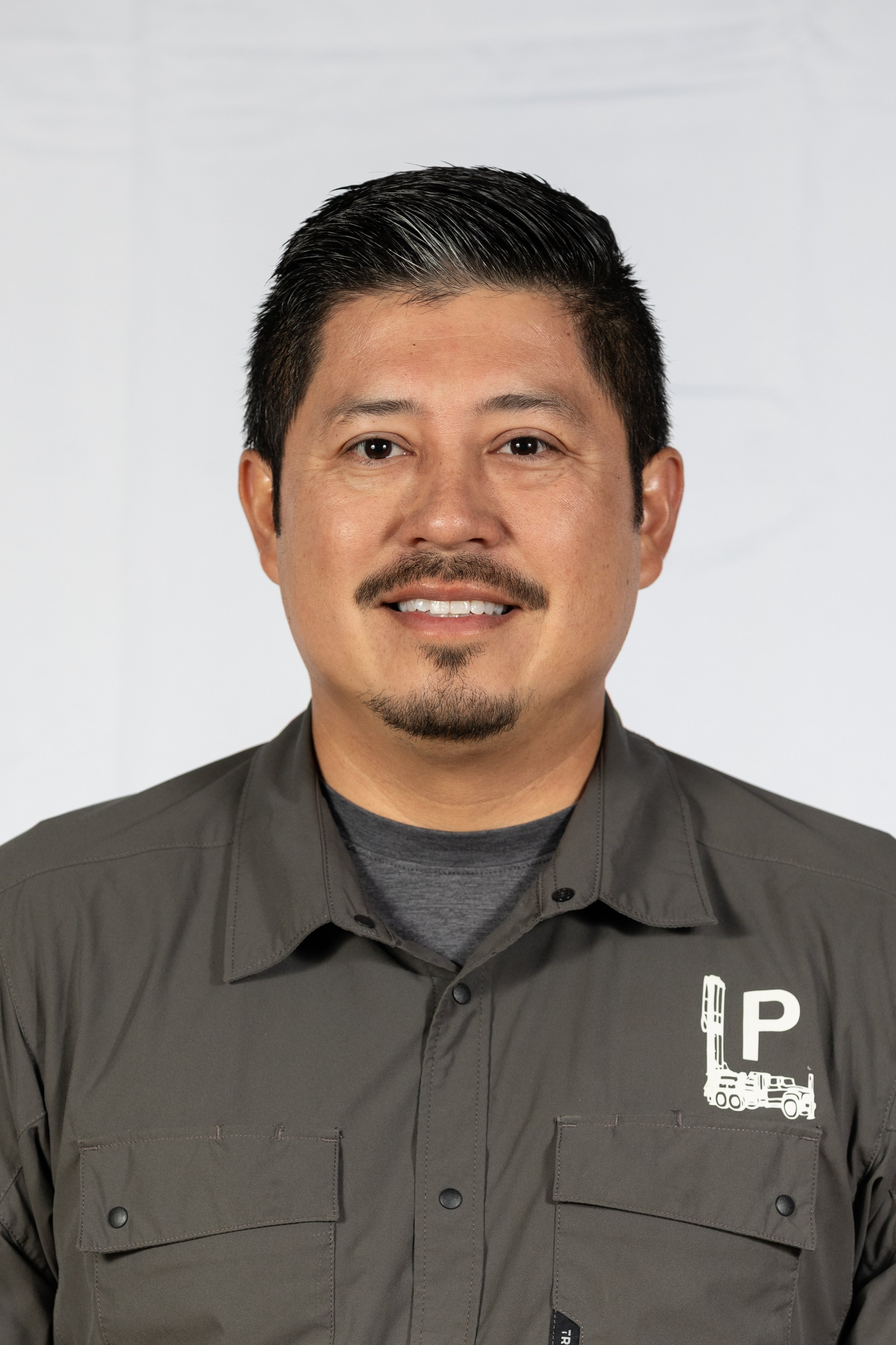 A man in a gray uniform with a drilling rig emblem on the chest, smiling in front of a plain background.