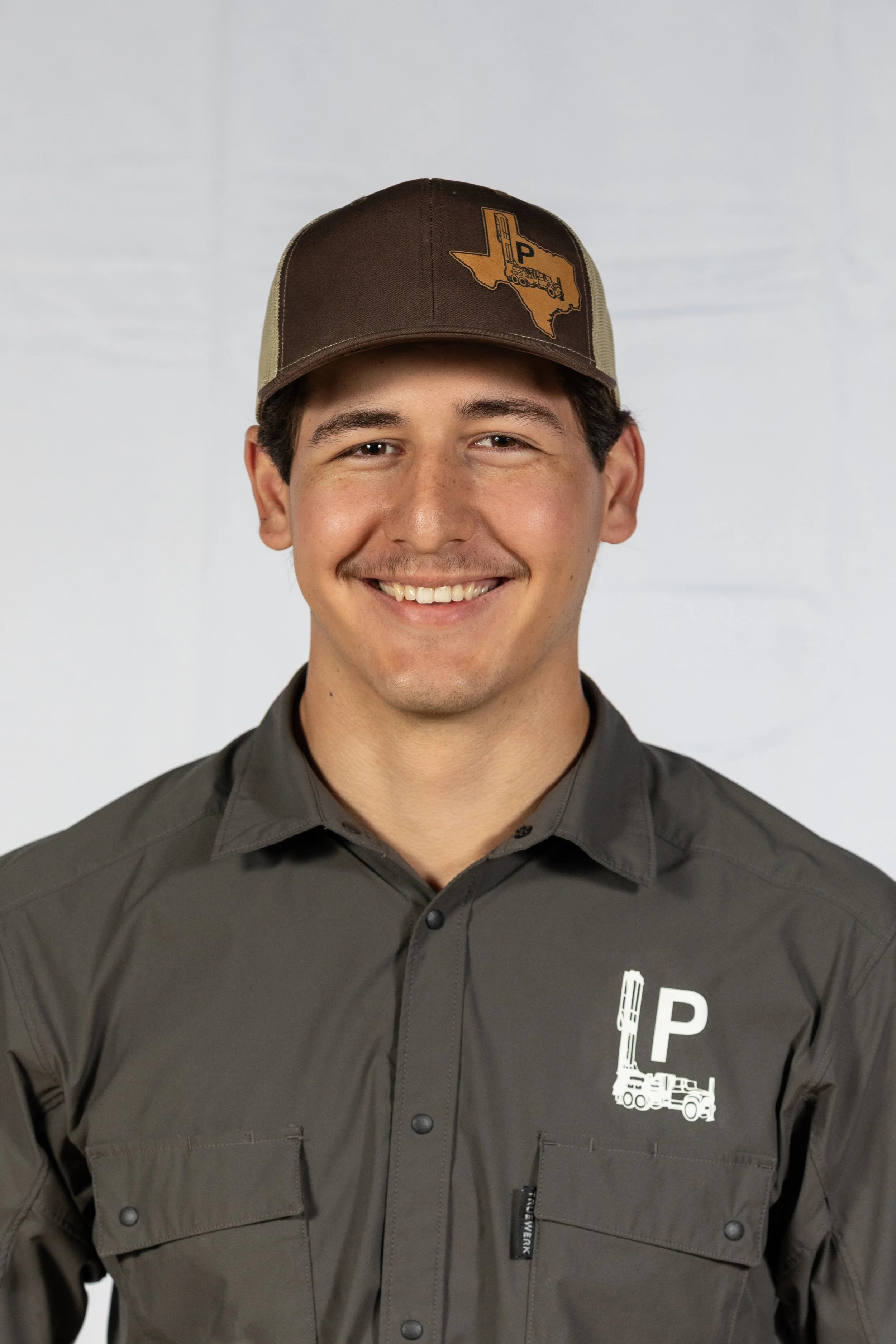 A young man smiling, wearing a brown cap with the outlines of Texas, and a gray button-up shirt with drilling equipment and a P logo on the shirt.