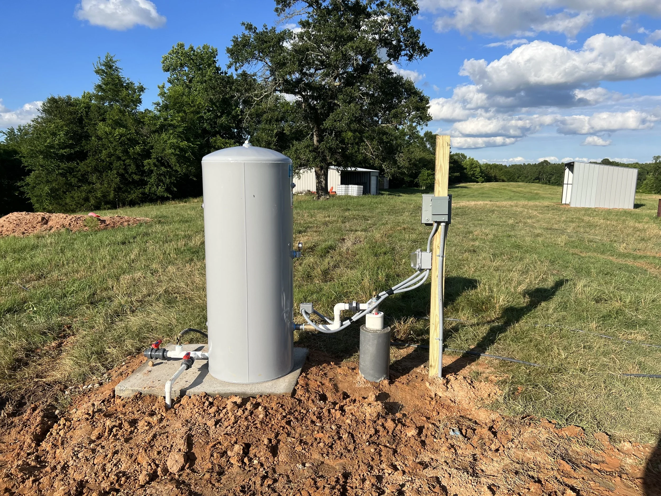 An outdoor setup with a tall, cylindrical white water tank connected to underground pipes, a wooden utility pole with electrical boxes, on grassy land under a partly cloudy sky, with trees and small metal buildings in the background.
