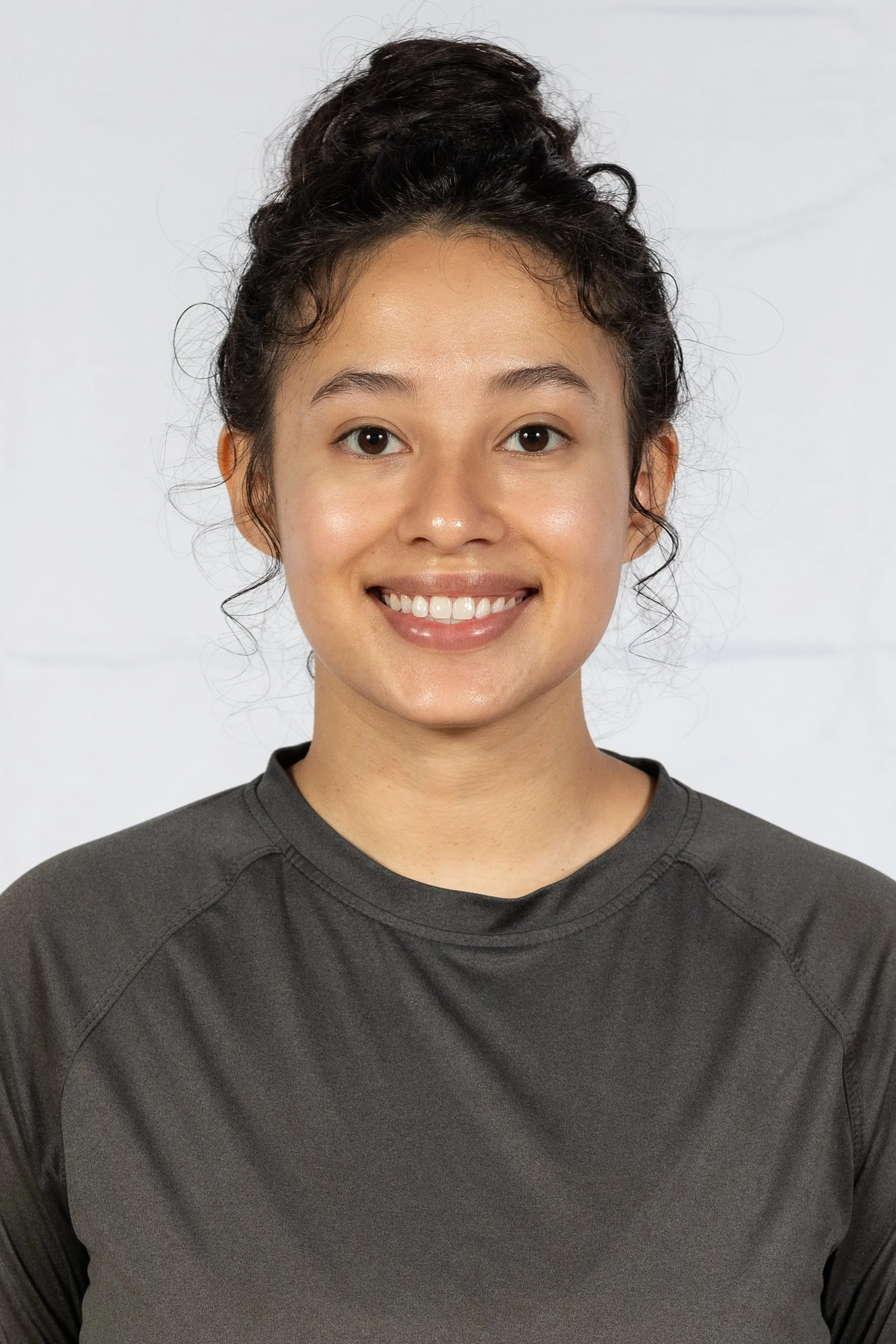 A young woman with dark hair styled in a messy bun, smiling, wearing a dark gray athletic shirt, in front of a plain white background.