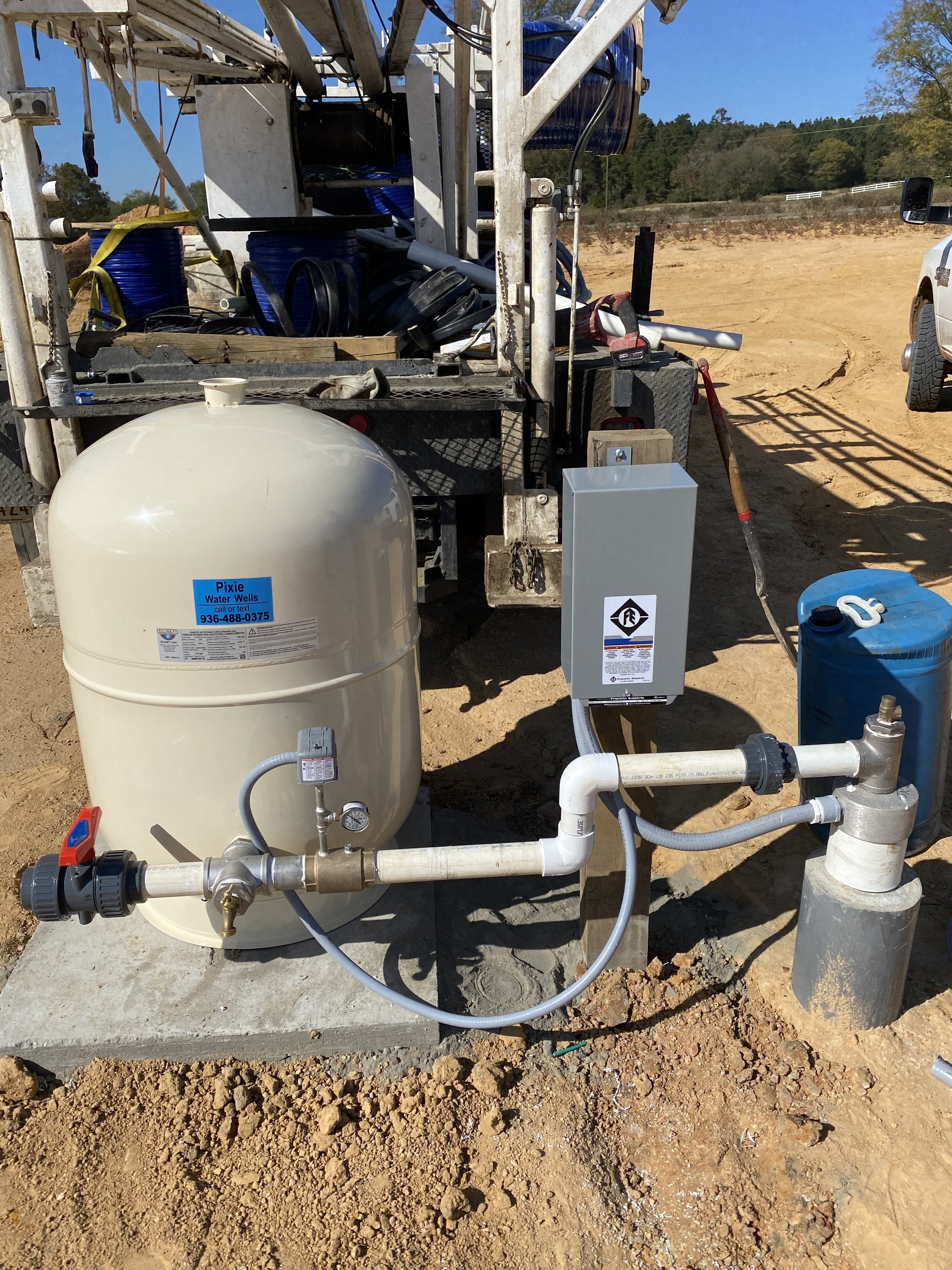 Water well equipment setup on a construction site, including a large white tank with a blue label, connected to pipes and valves, with a pulling unit and a rural landscape in the background.