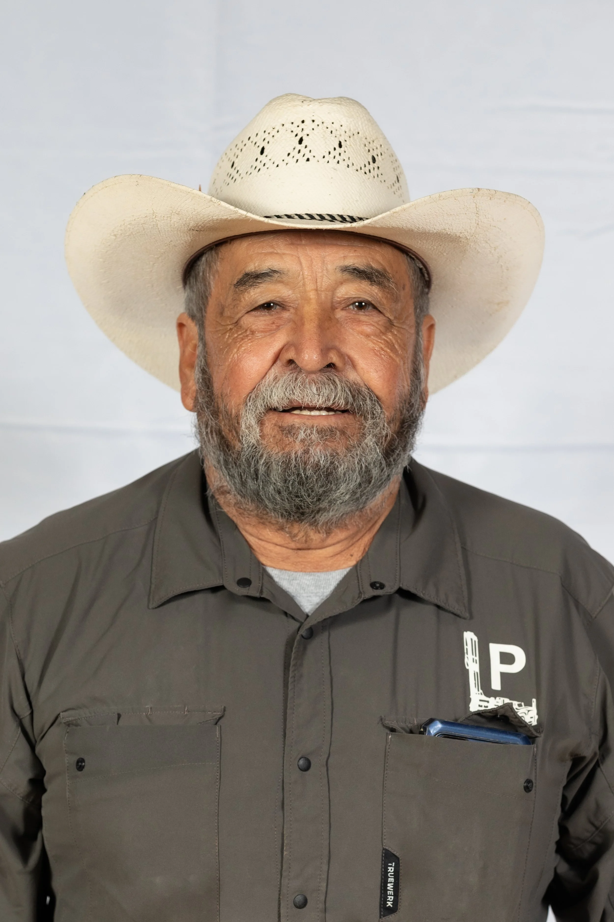 Older bearded man wearing a light straw cowboy hat and a dark button-up shirt with a white drilling rig and letter 'P' logo on the chest, standing against a white background.