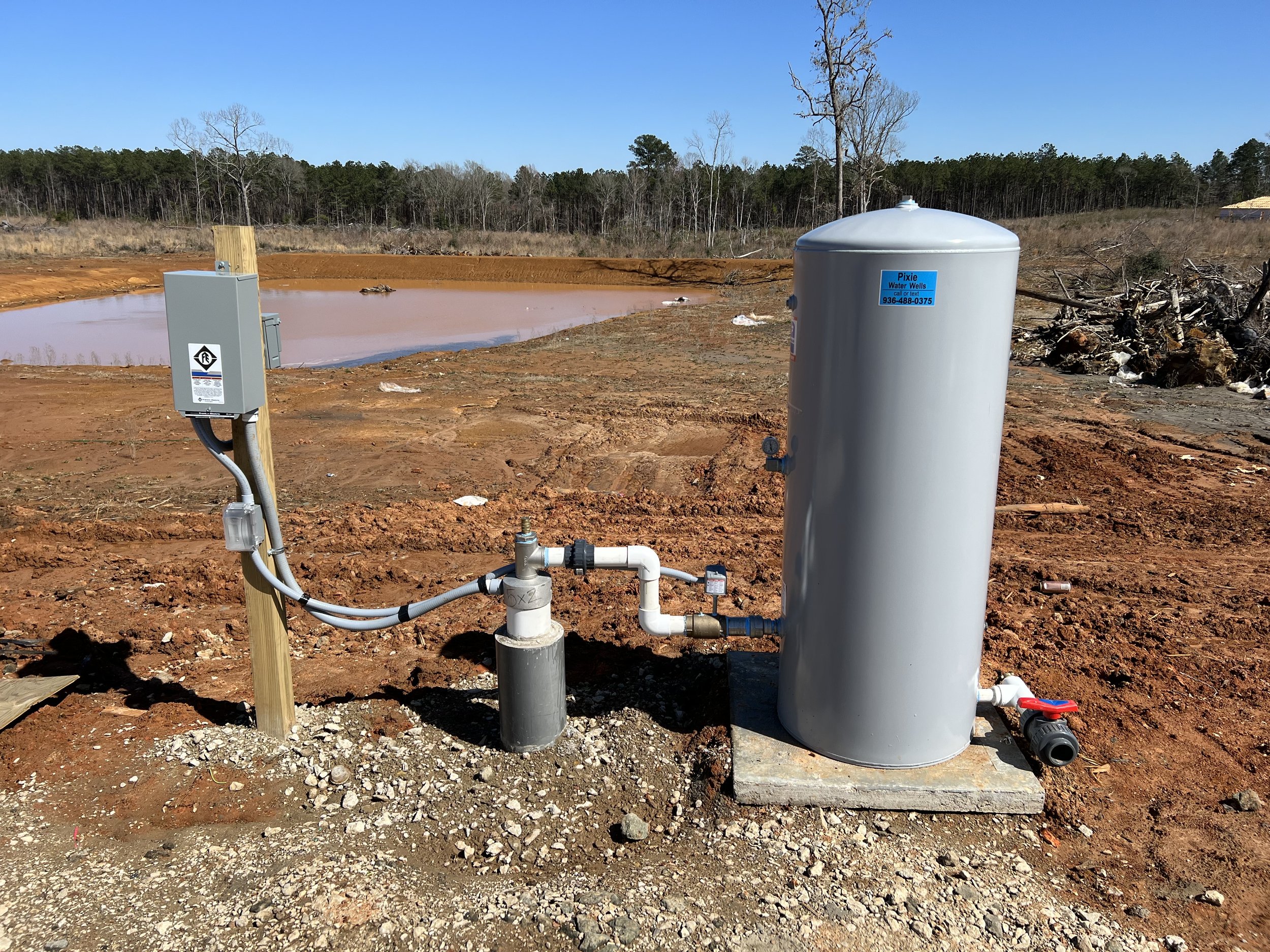 Water well system with electrical panel on wooden post and a large grey cylindrical tank, situated on a concrete slab near a pond and rural landscape.