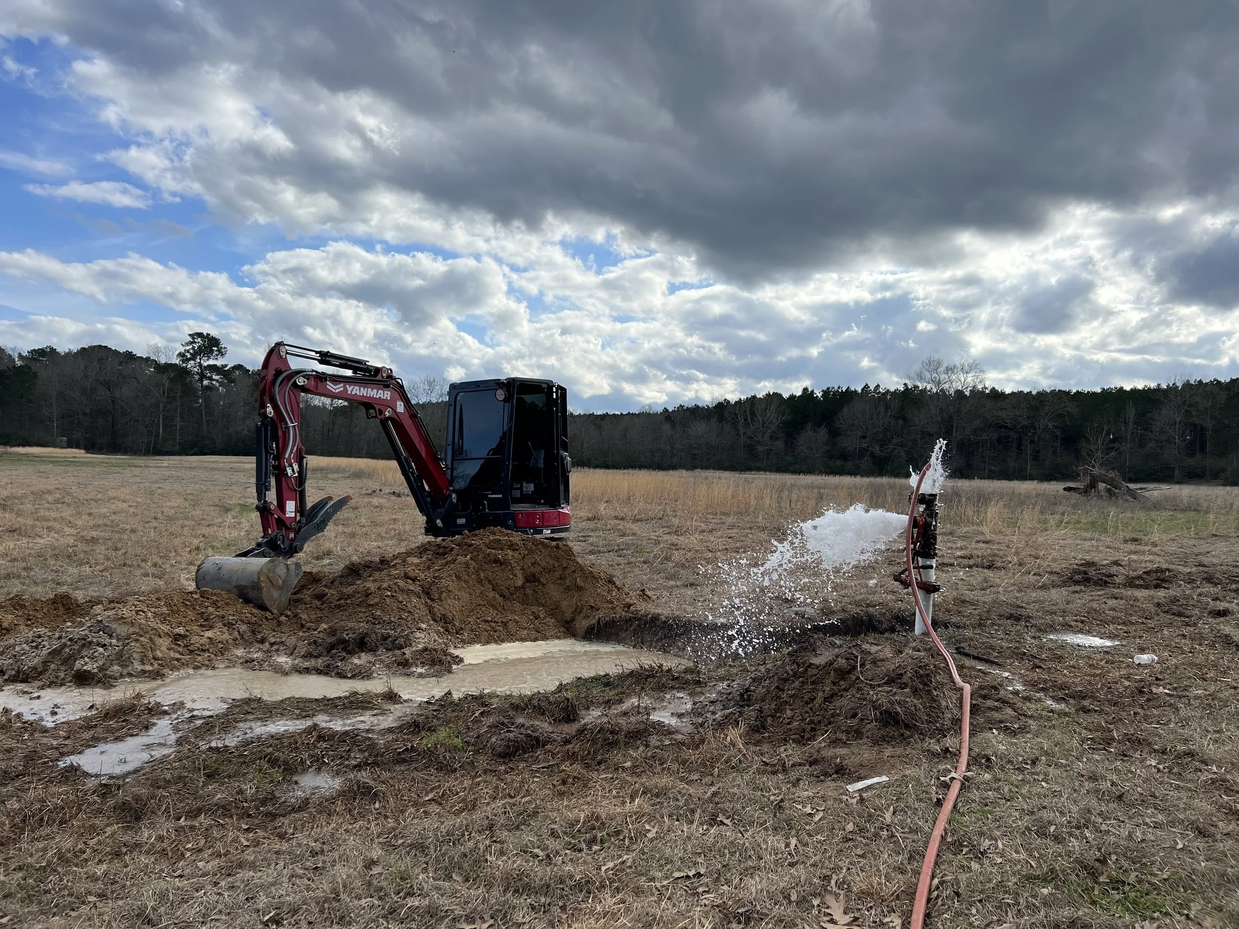 A small red and black excavator digging in an open field with cloudy sky overhead.