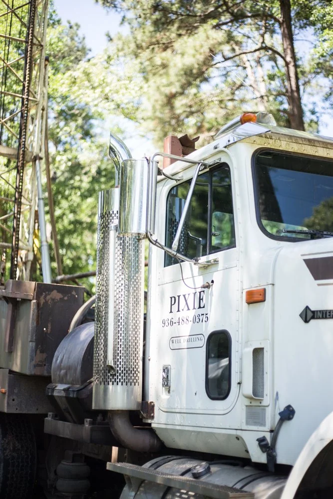 Close-up of a white drilling rig labeled "Pixie" with a phone number, parked outdoors with trees in the background.