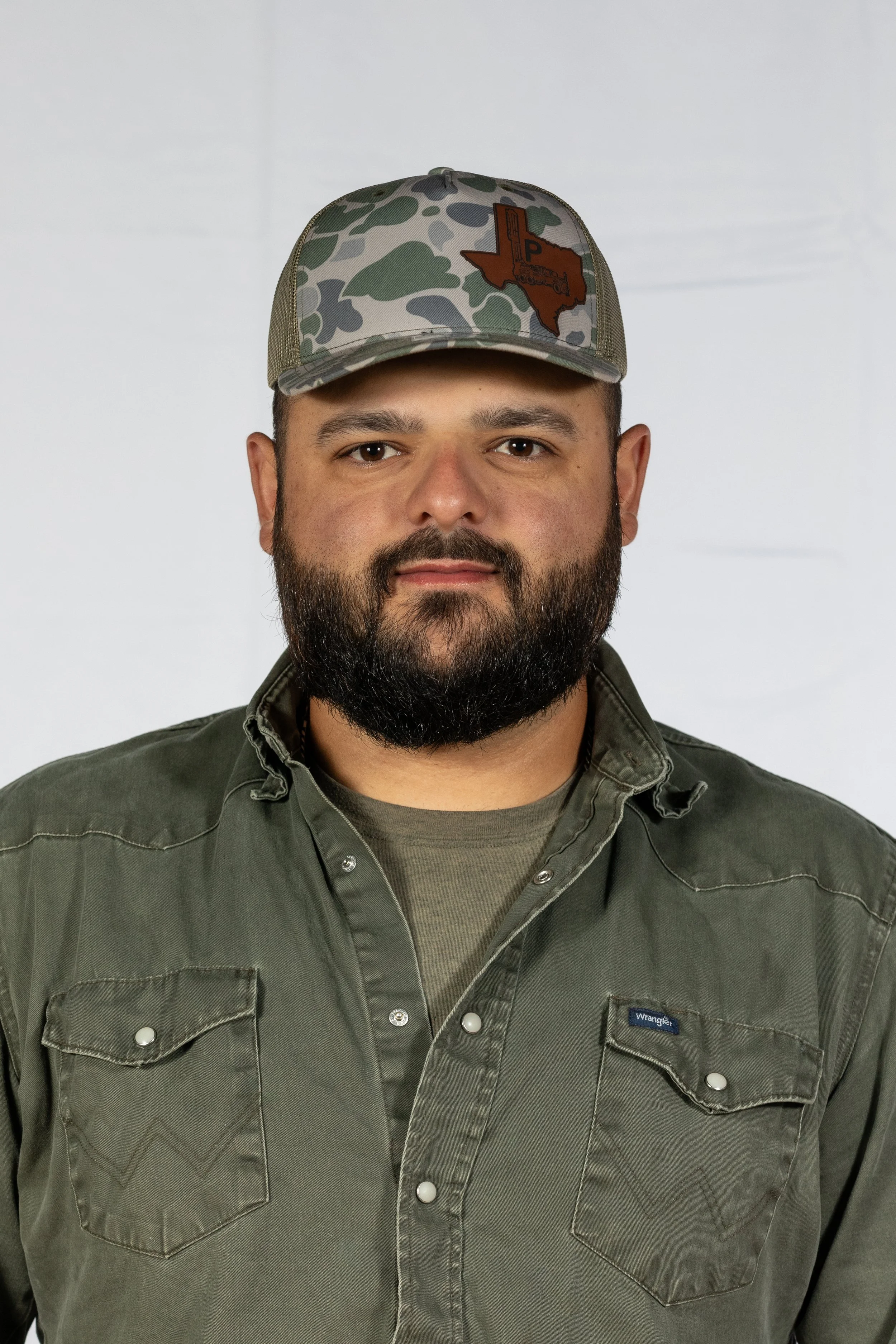 A man with a beard wearing a green button-up shirt and a camouflage cap with a Texas-shaped patch.