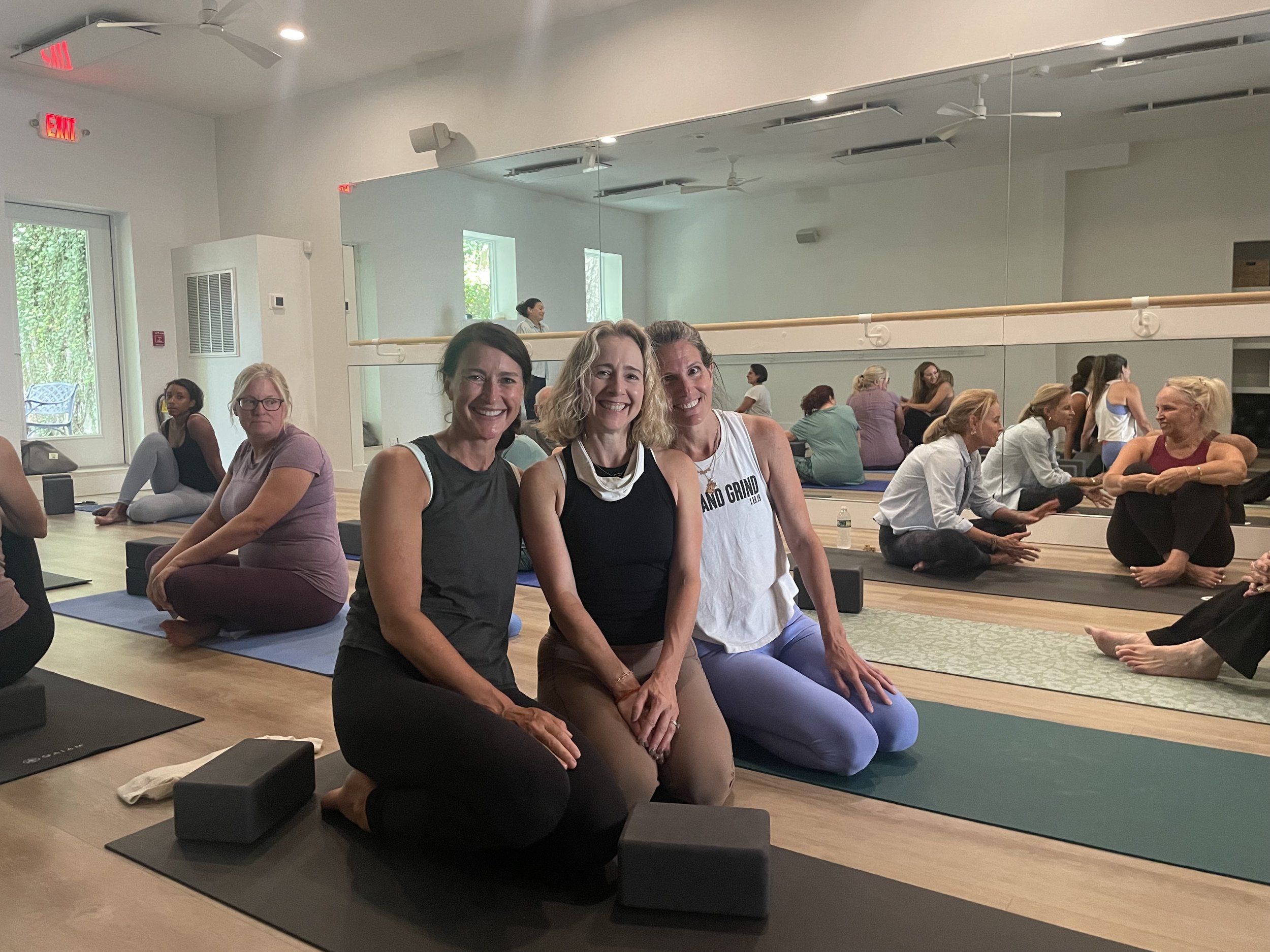 Three women kneeling on yoga mats smiling in a yoga studio, with other women practicing yoga in the background.