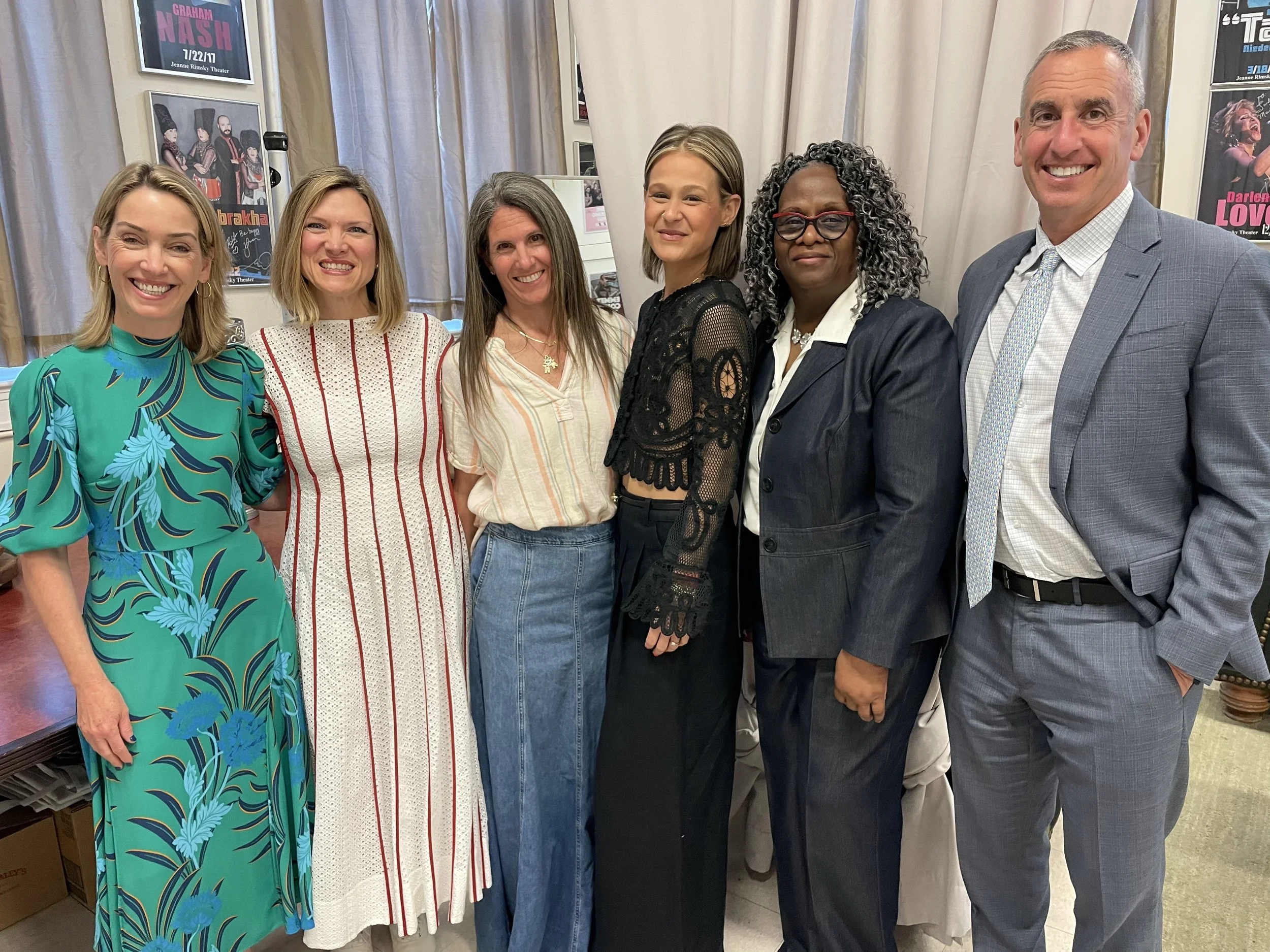 Six people standing indoors, posing for a photo. They are smiling and dressed in various colorful and professional outfits. The background shows posters on the wall.