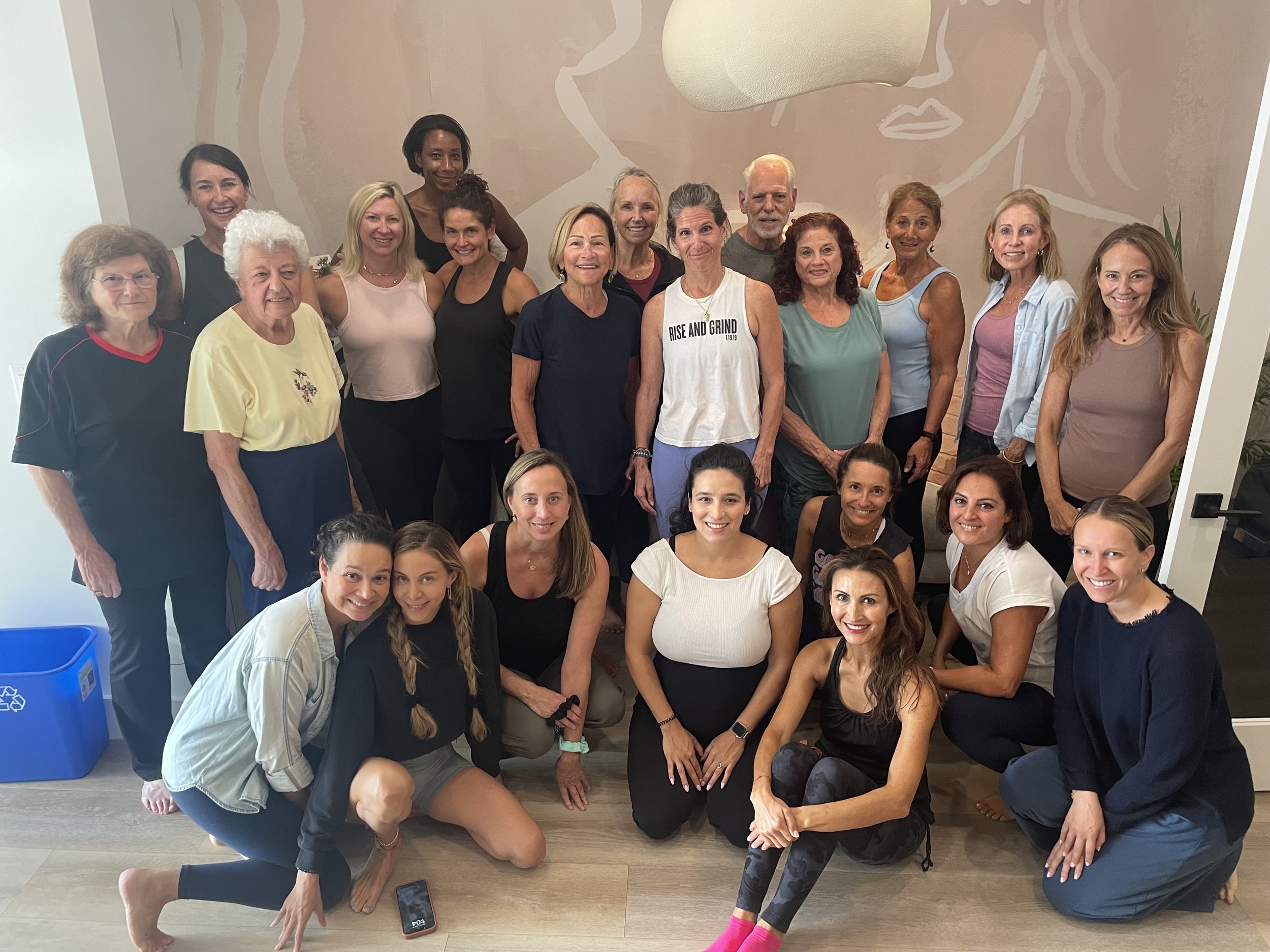 A group of 20 women of various ages posing together indoors, some standing and some sitting in front, smiling and looking at the camera.