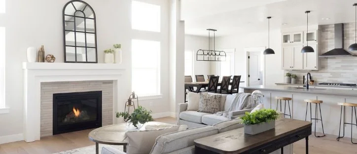 Bright, open living room and kitchen with white walls, a fireplace, sofa, and a dining area with dark chairs and a kitchen with white cabinets and black pendant lights.