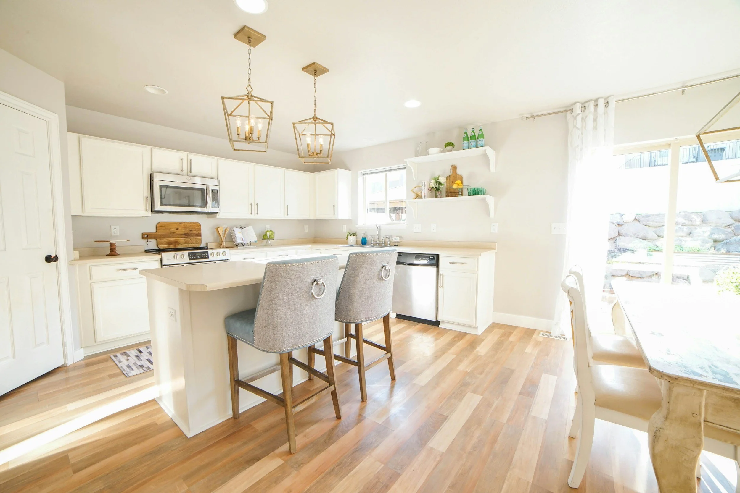 Bright kitchen with white cabinets, a kitchen island, two gray barstools, a dining table with white chairs, overhead lighting, hardwood floors, and open shelving with decorative items, with sunlight coming through a sliding glass door.
