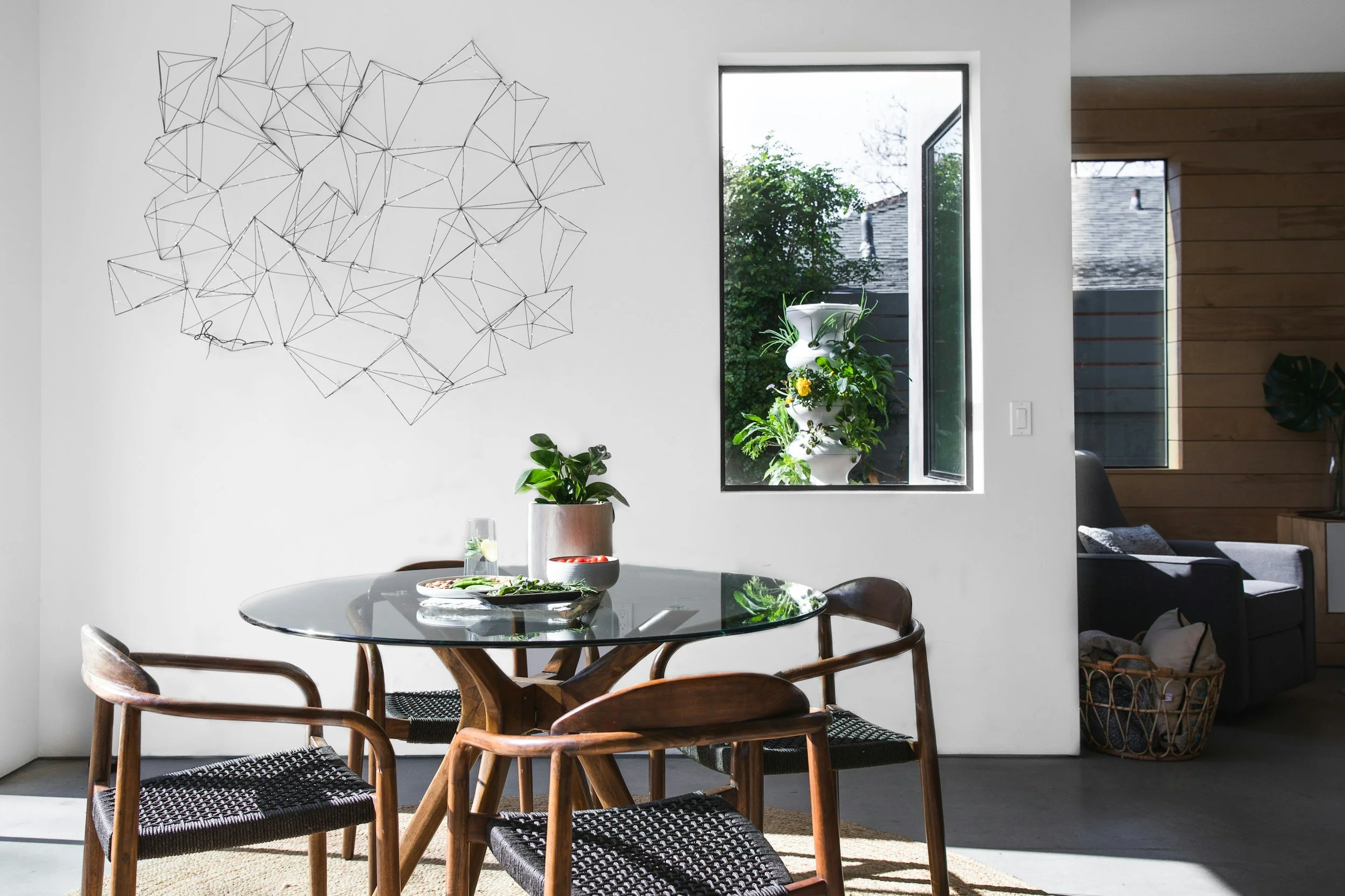 Modern dining area with a glass-topped round table, four wooden chairs with woven seats, and indoor plants. A large wall art piece made of geometric wire shapes is on the white wall. Sunlight streams in through an open window, revealing a view of greenery outside. In the background, a living room with wooden paneling, a gray armchair, and a basket of blankets is visible.